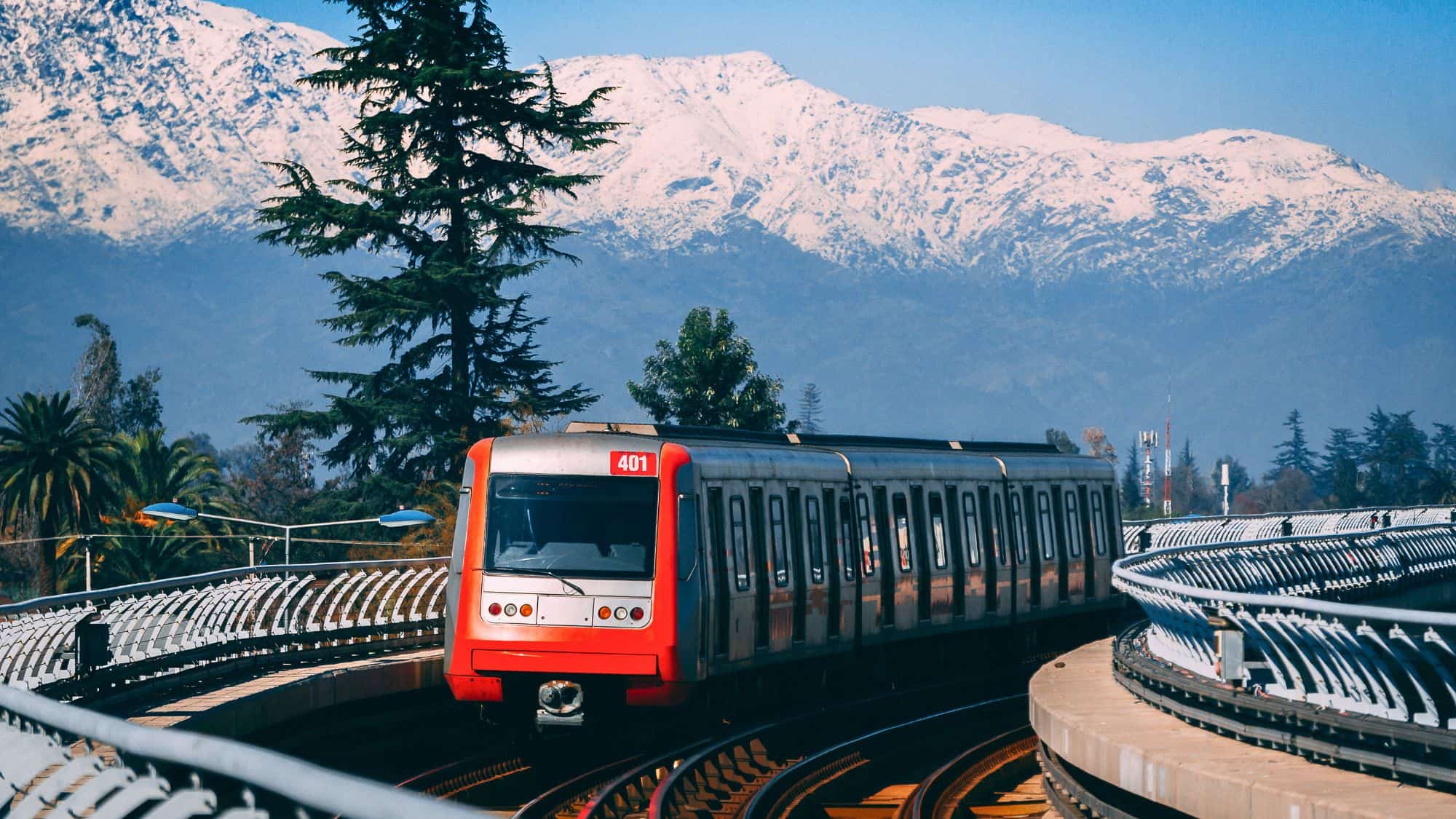 A modern red metro train travels along elevated tracks with snow-capped mountains in the background.