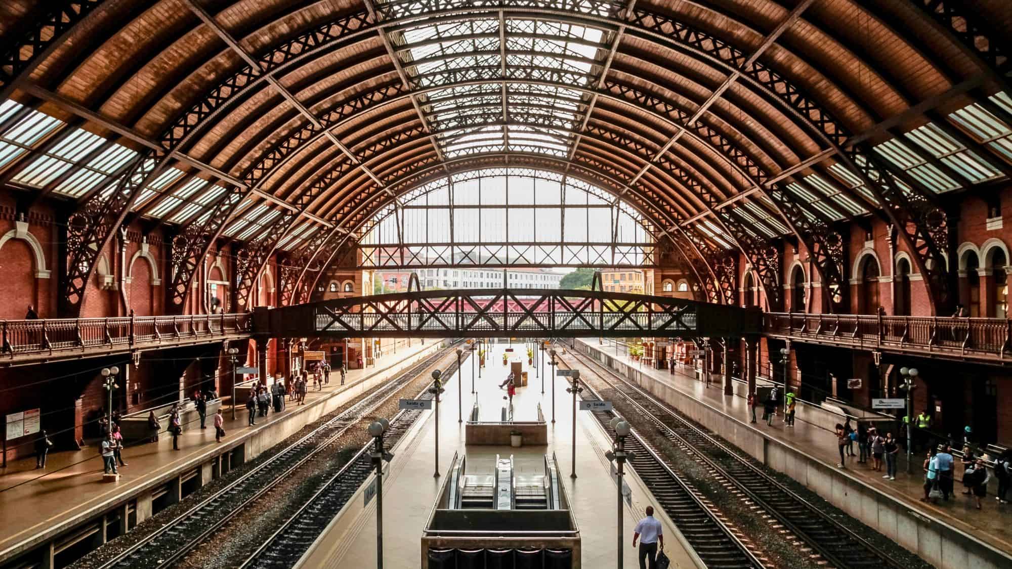 A wide view of a grand train station interior with arched ceilings, iron beams, and multiple tracks lined with passengers.