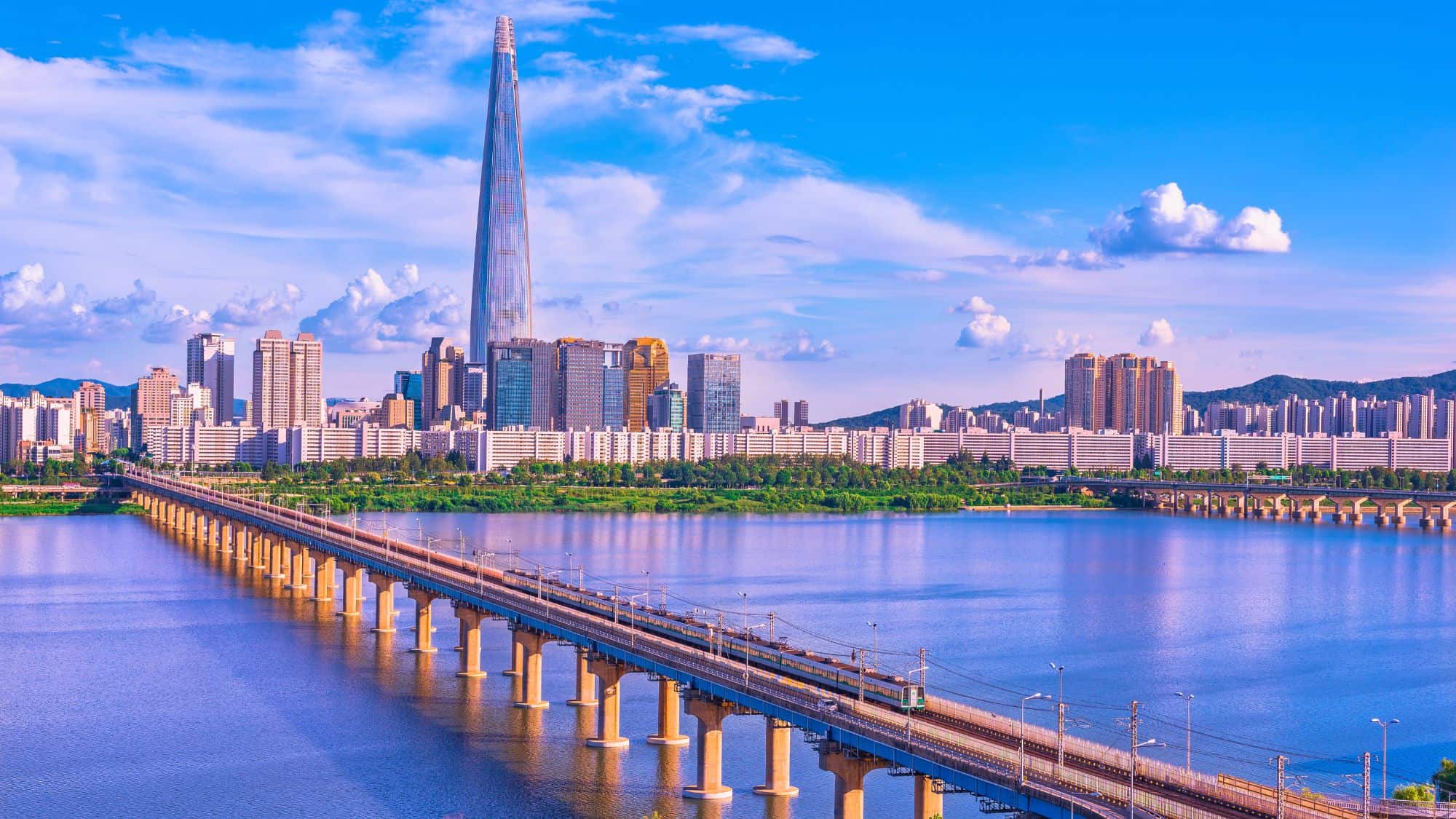 A commuter train runs across a long bridge over the Han River with Seoul’s skyline and Lotte World Tower rising in the distance.