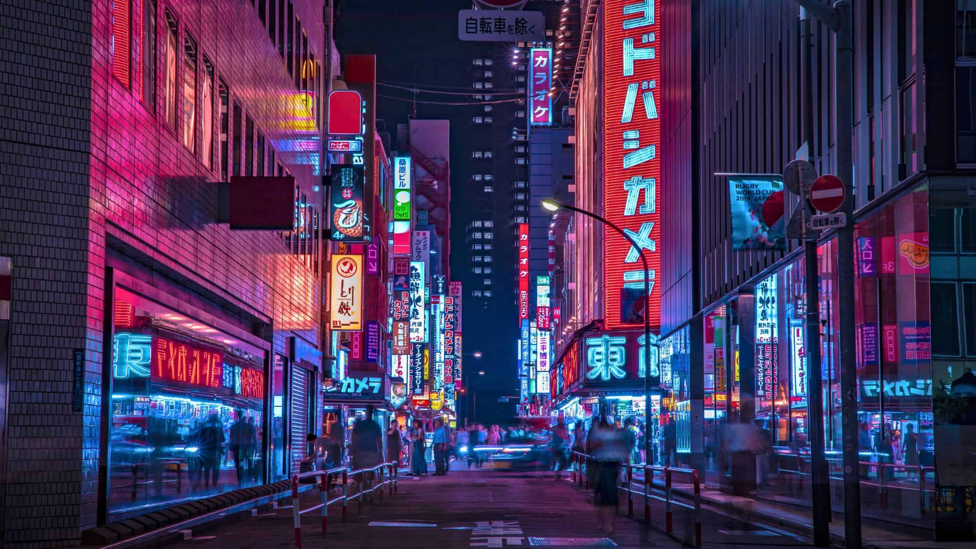 Tokyo's Shinjuku district bursts with towering neon signs in Japanese and glowing storefronts, as blurred figures bustle down a narrow street after dark.