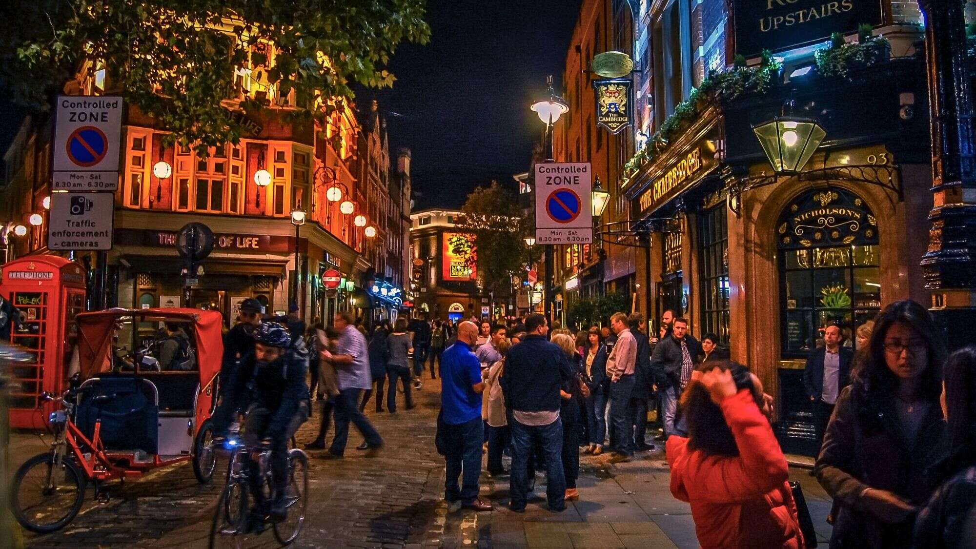 People gather outside classic English pubs on a cobblestone street in London’s Soho, with red phone booths, pub signs, and a cyclist weaving through.