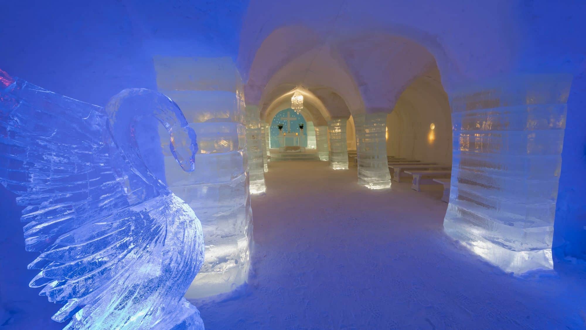 A frosty interior with thick ice columns and benches leads to an altar with a glowing blue cross, with a detailed ice sculpture of a swan prominently displayed in the foreground.