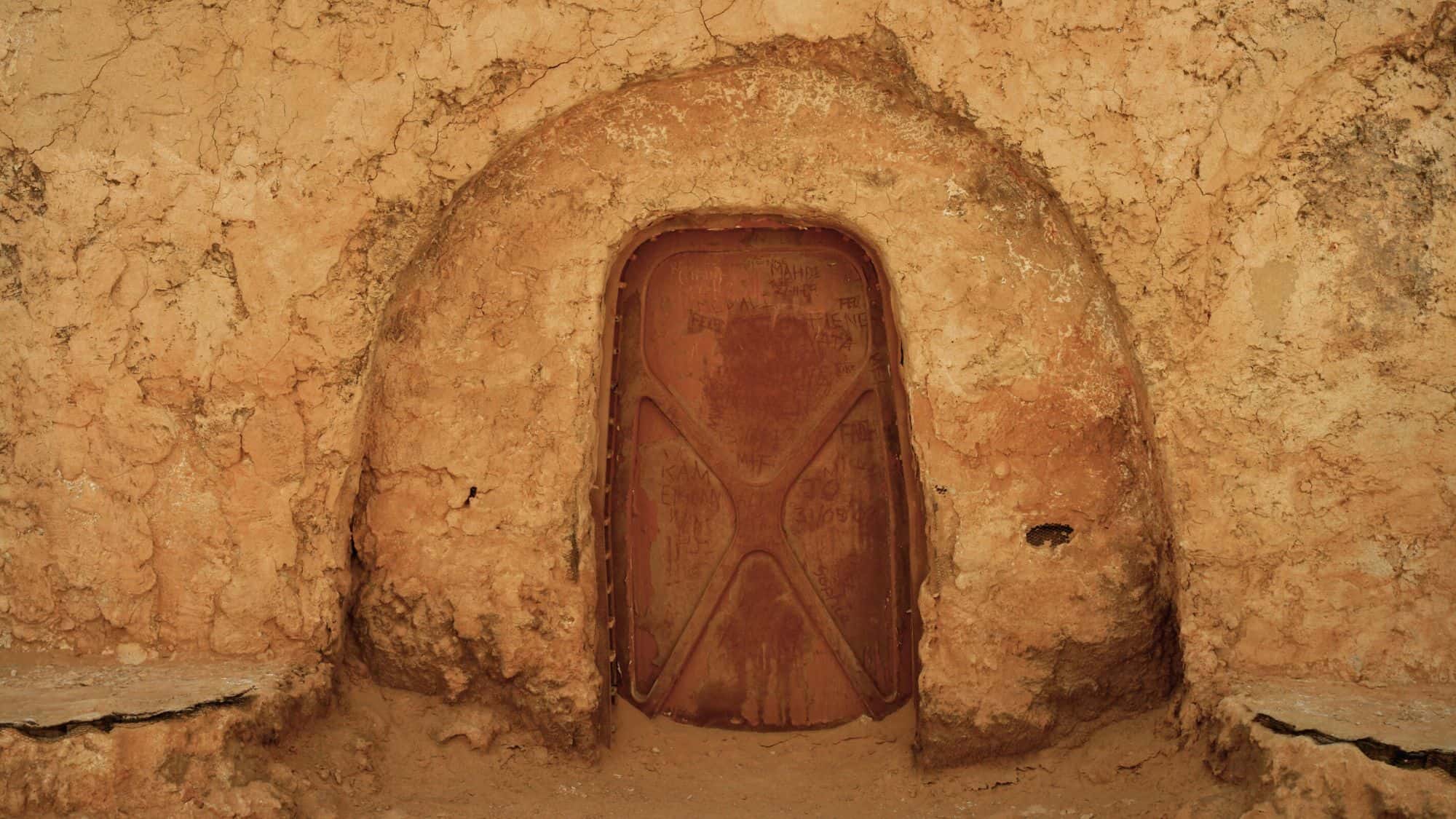 A weathered, rust-colored metal door is set into a sandy, earthen wall with a rounded arch, part of the abandoned Star Wars film set in Matmata, Tunisia, representing Luke Skywalker's childhood home on Tatooine.