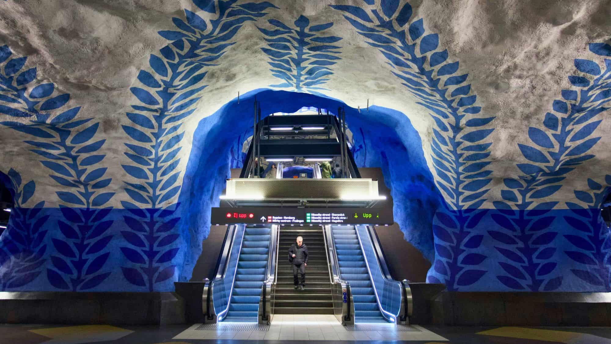 An escalator leads up from a Stockholm subway station, where blue leaf patterns are painted on the cave-like walls.