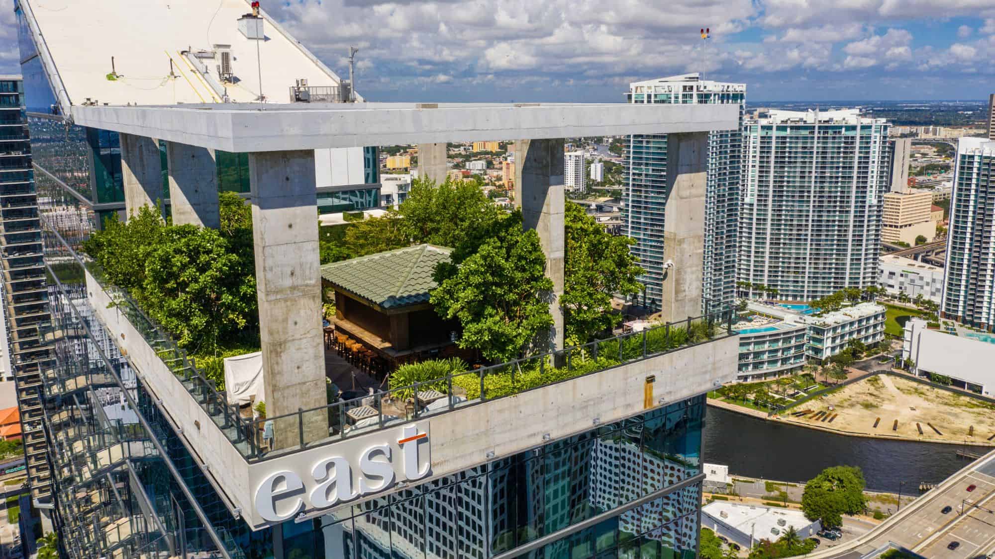 A sleek rooftop bar filled with lush tropical trees and minimalist design sits atop a high-rise in Miami, enclosed by concrete columns and surrounded by tall residential towers.