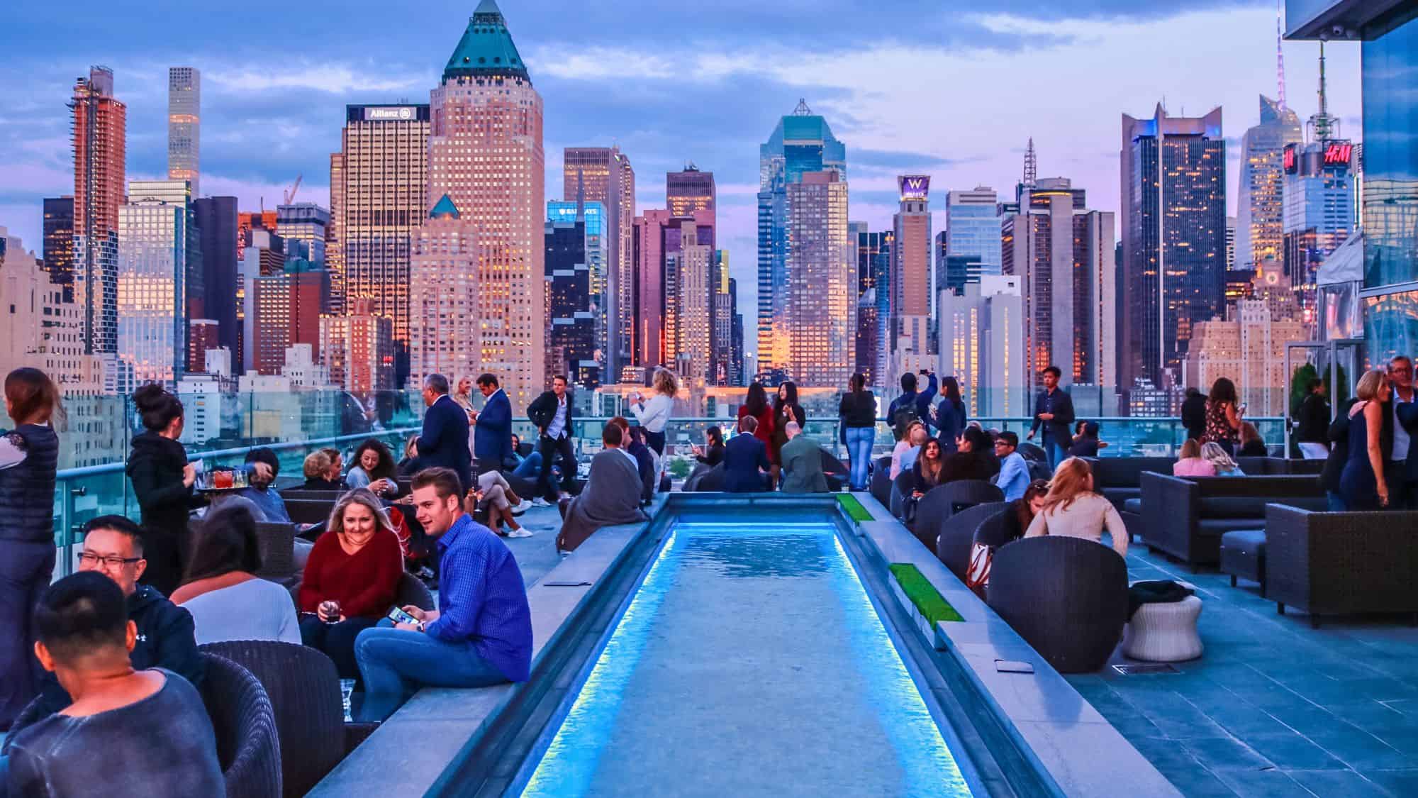 A lively rooftop bar with a narrow illuminated reflecting pool is packed with people enjoying cocktails and city views, with Manhattan’s colorful skyline glowing at dusk in the background.