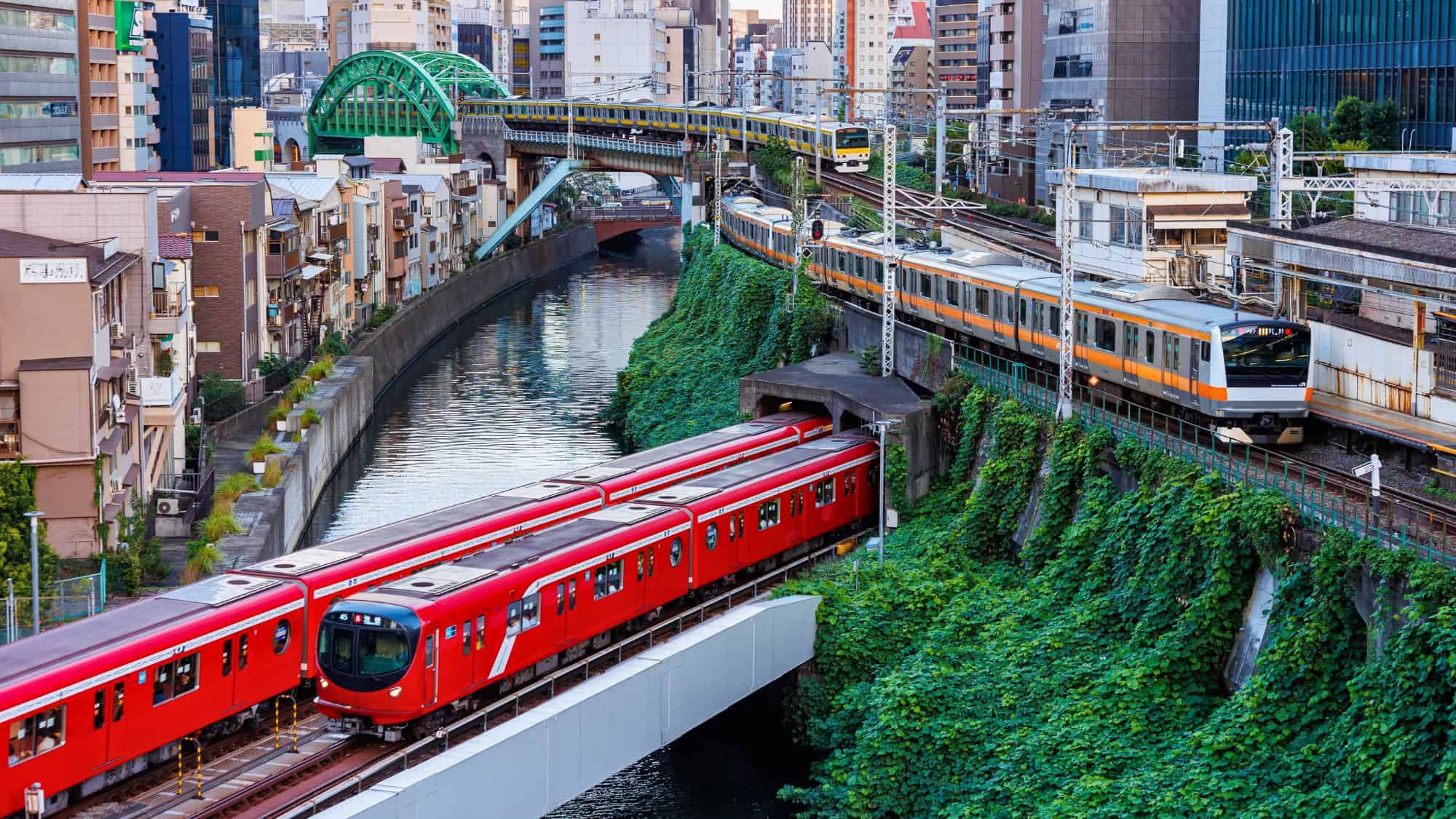 Multiple trains, including bright red and orange carriages, run along elevated tracks above a canal lined with greenery and city buildings in Tokyo.
