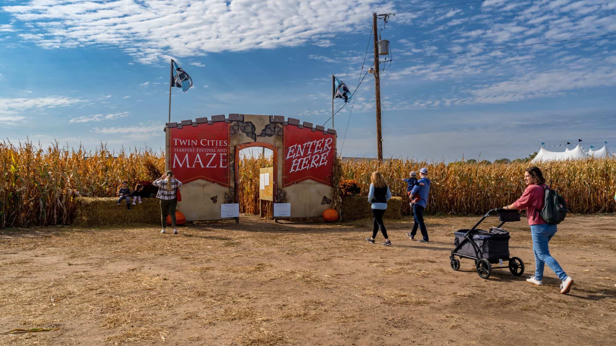 A castle-themed entrance labeled “Twin Cities Harvest Festival and Maze” welcomes families with children and strollers into a corn maze on a sunny fall day.