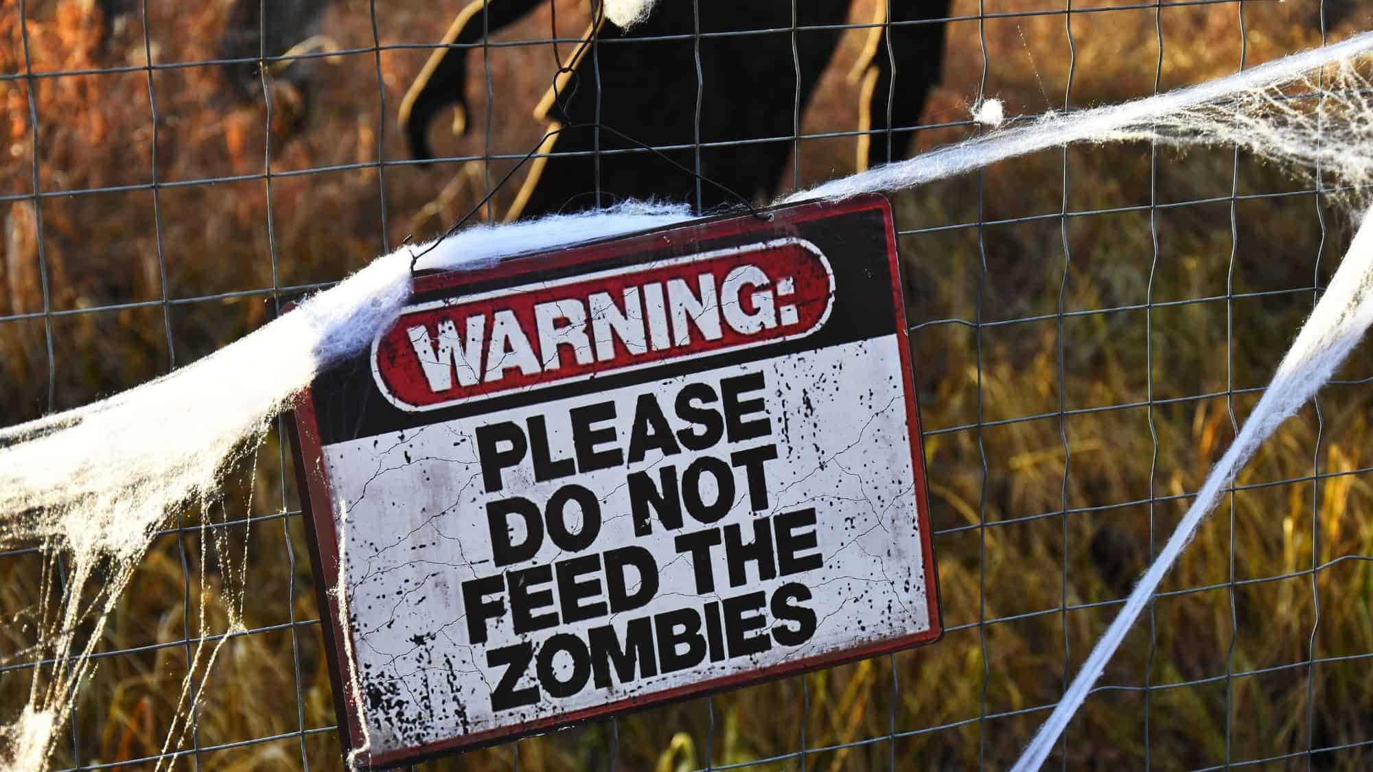 A distressed metal sign on a wire fence reads "WARNING: PLEASE DO NOT FEED THE ZOMBIES," surrounded by fake cobwebs in a dry outdoor setting.
