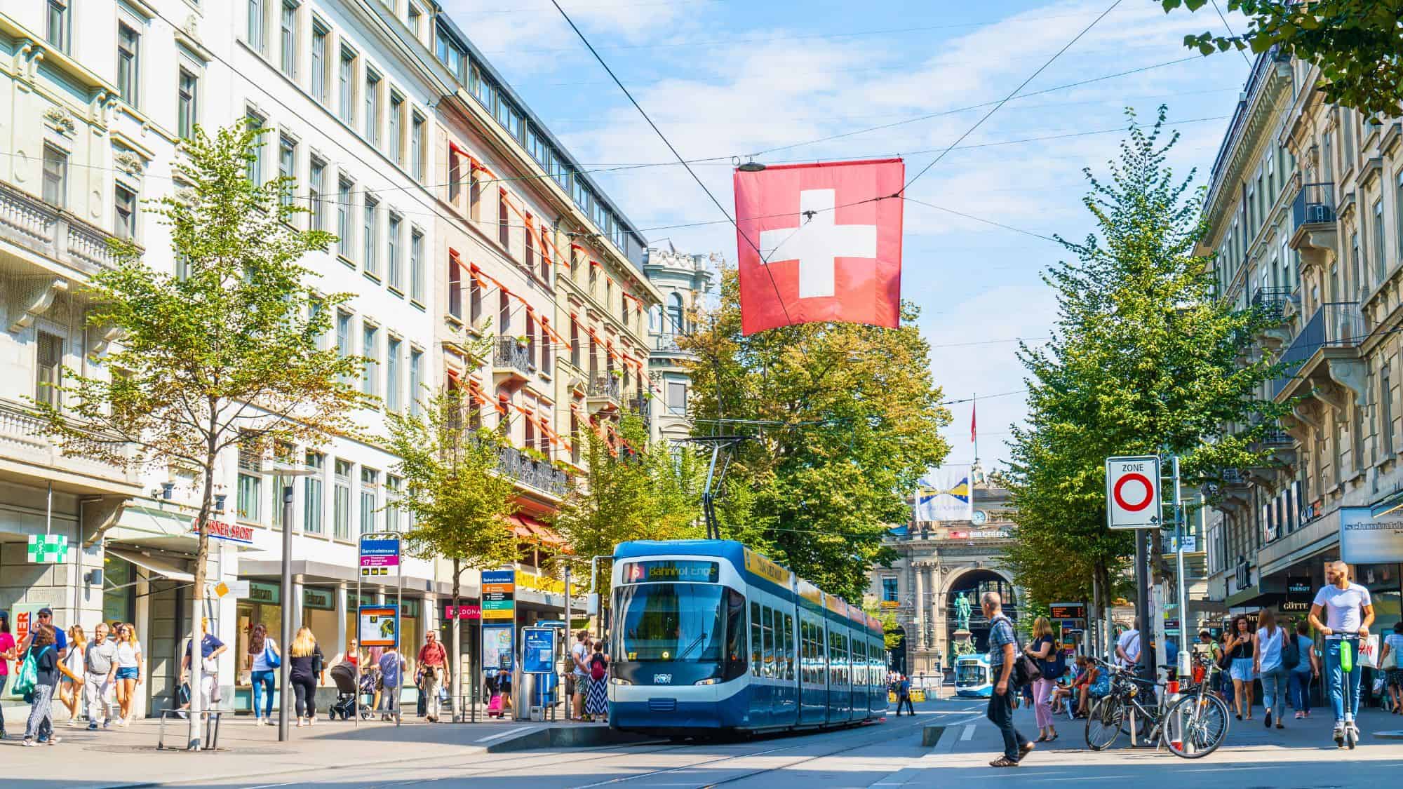 A blue tram moves through a busy Zurich street lined with historic buildings, with a Swiss flag hanging above.