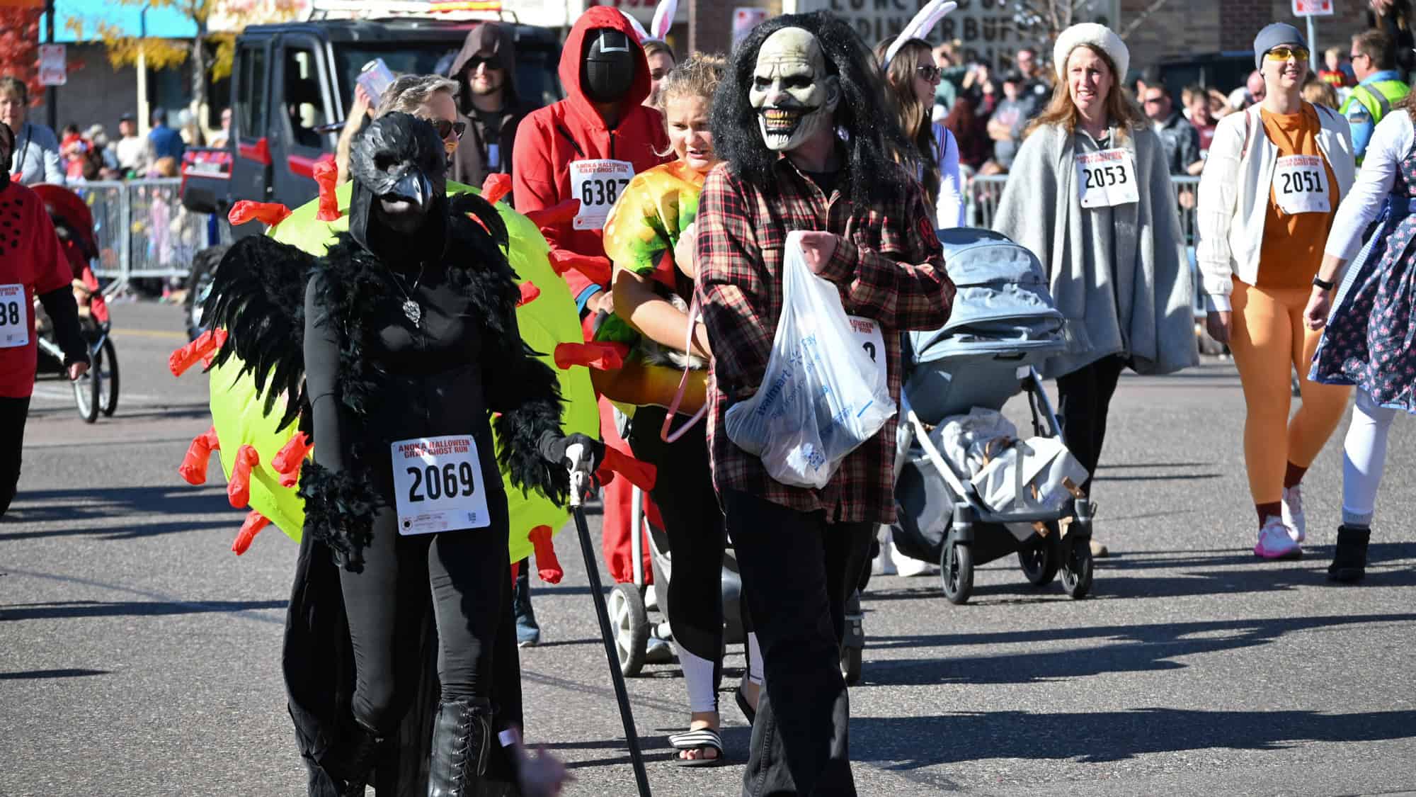 Participants in a Halloween-themed fun run walk in colorful costumes, including a plague crow, monster mask, and food-themed outfits, all wearing numbered race bibs.