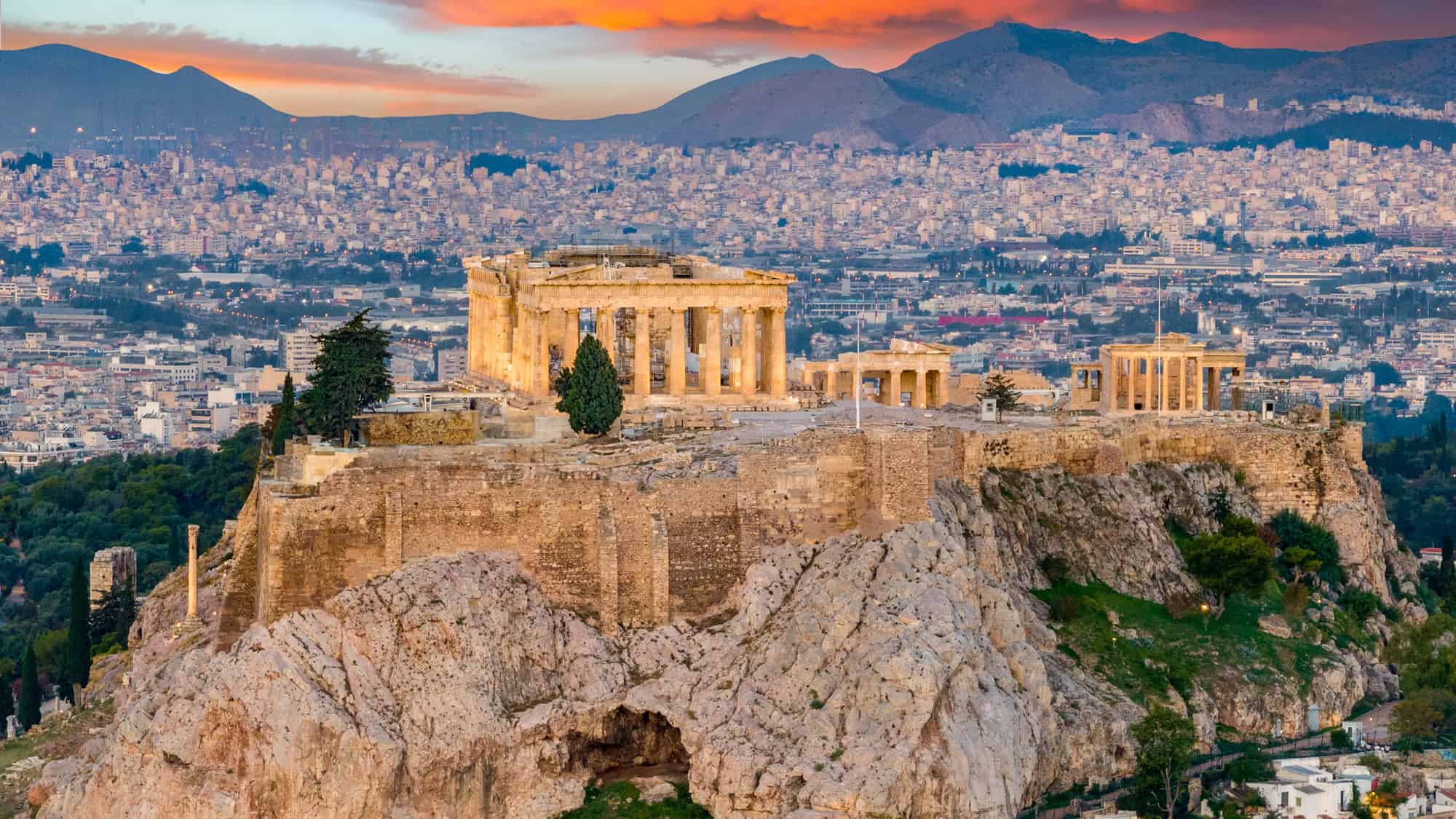 The ancient ruins of the Acropolis in Athens, Greece stand illuminated atop a rocky hill, with a dense cityscape and dramatic sunset sky in the background.