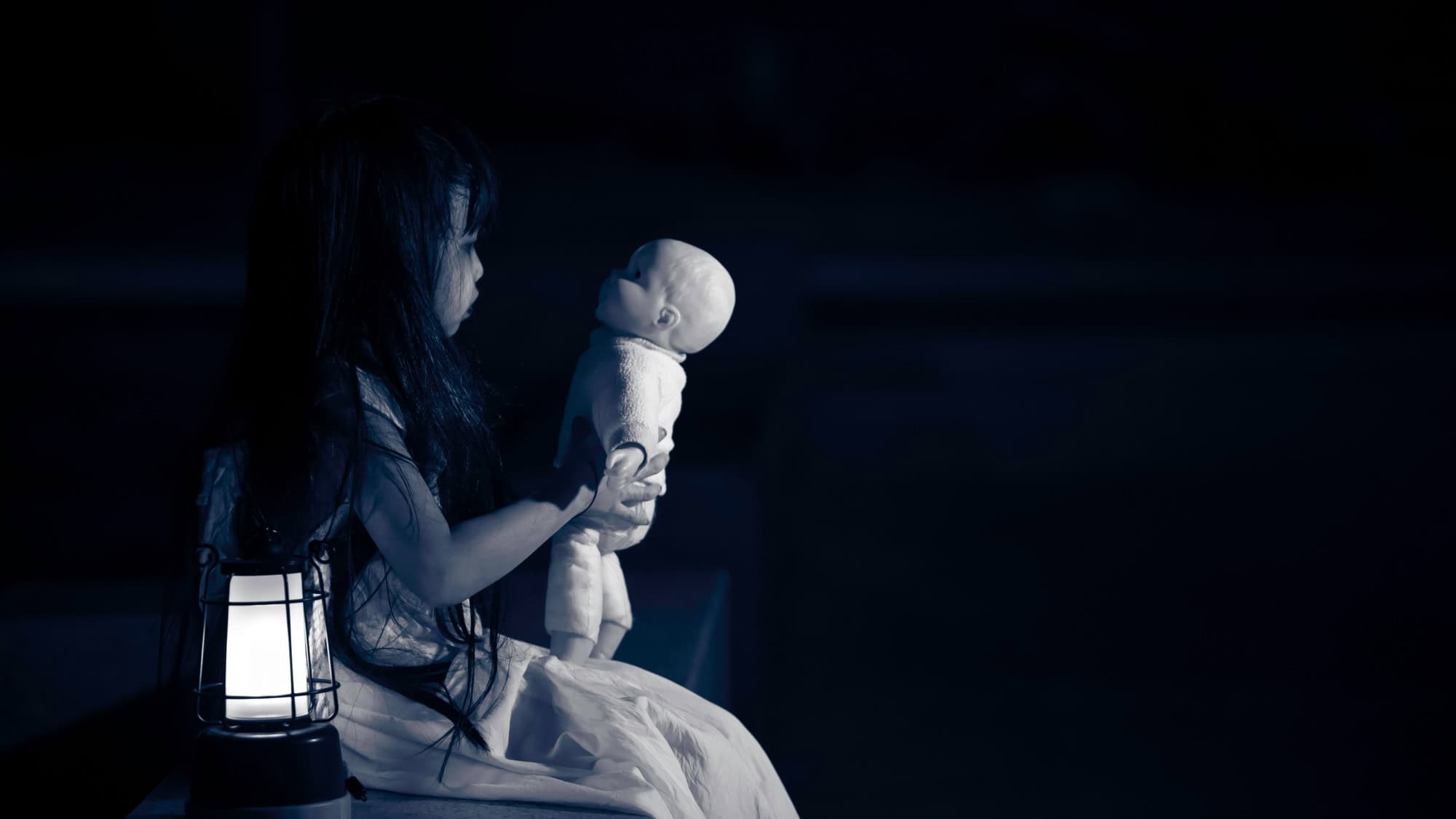 In dim blue light, a long-haired doll in a white dress eerily holds a baby doll while sitting beside a lantern, creating an unsettling and ghostly atmosphere.