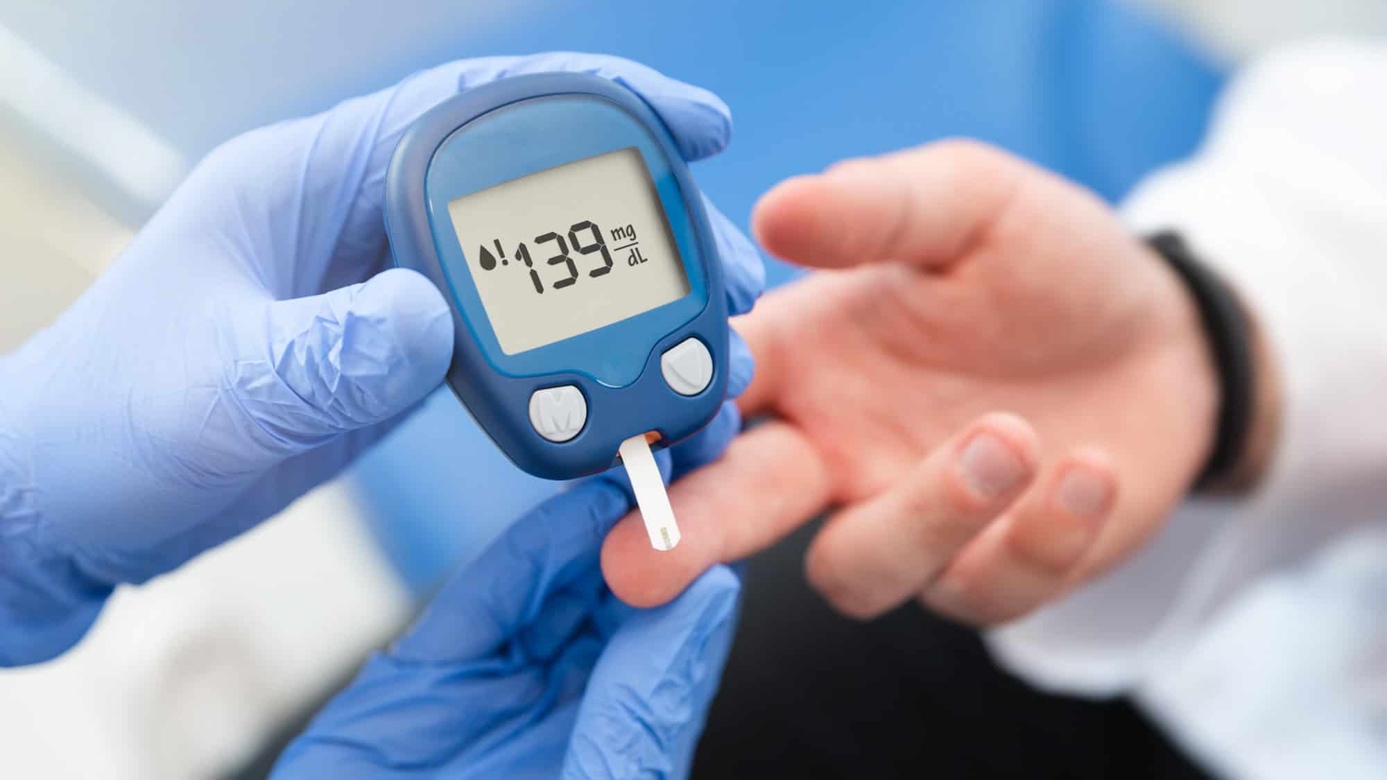 A gloved hand uses a glucose meter showing a reading of 139 mg/dL as another person’s finger is tested.