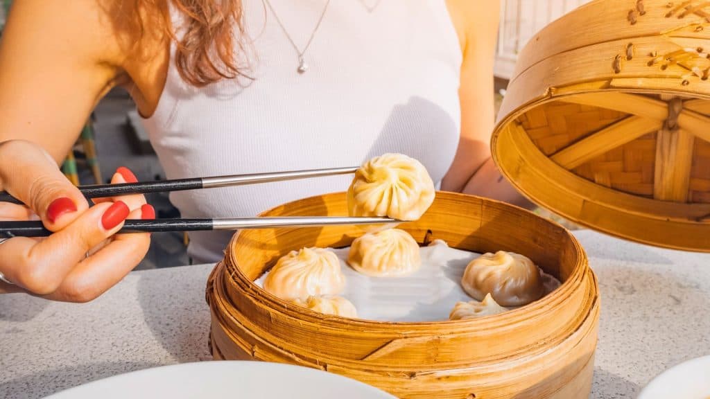 A woman in a white tank top lifts a soup dumpling with chopsticks from a bamboo steamer filled with neatly arranged xiao long bao.