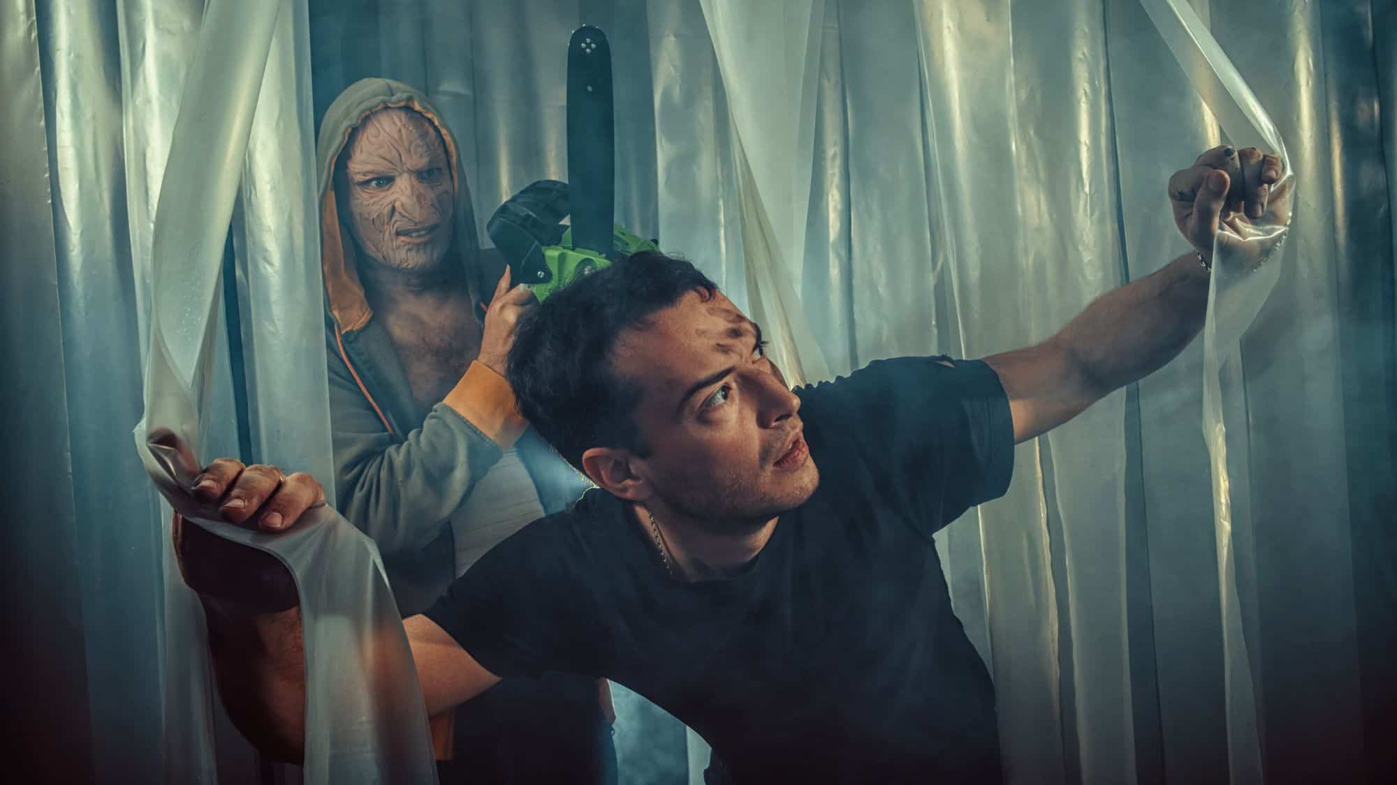 A terrified man pushes through plastic sheeting unaware that a masked killer holding a chainsaw is silently stalking him from behind.