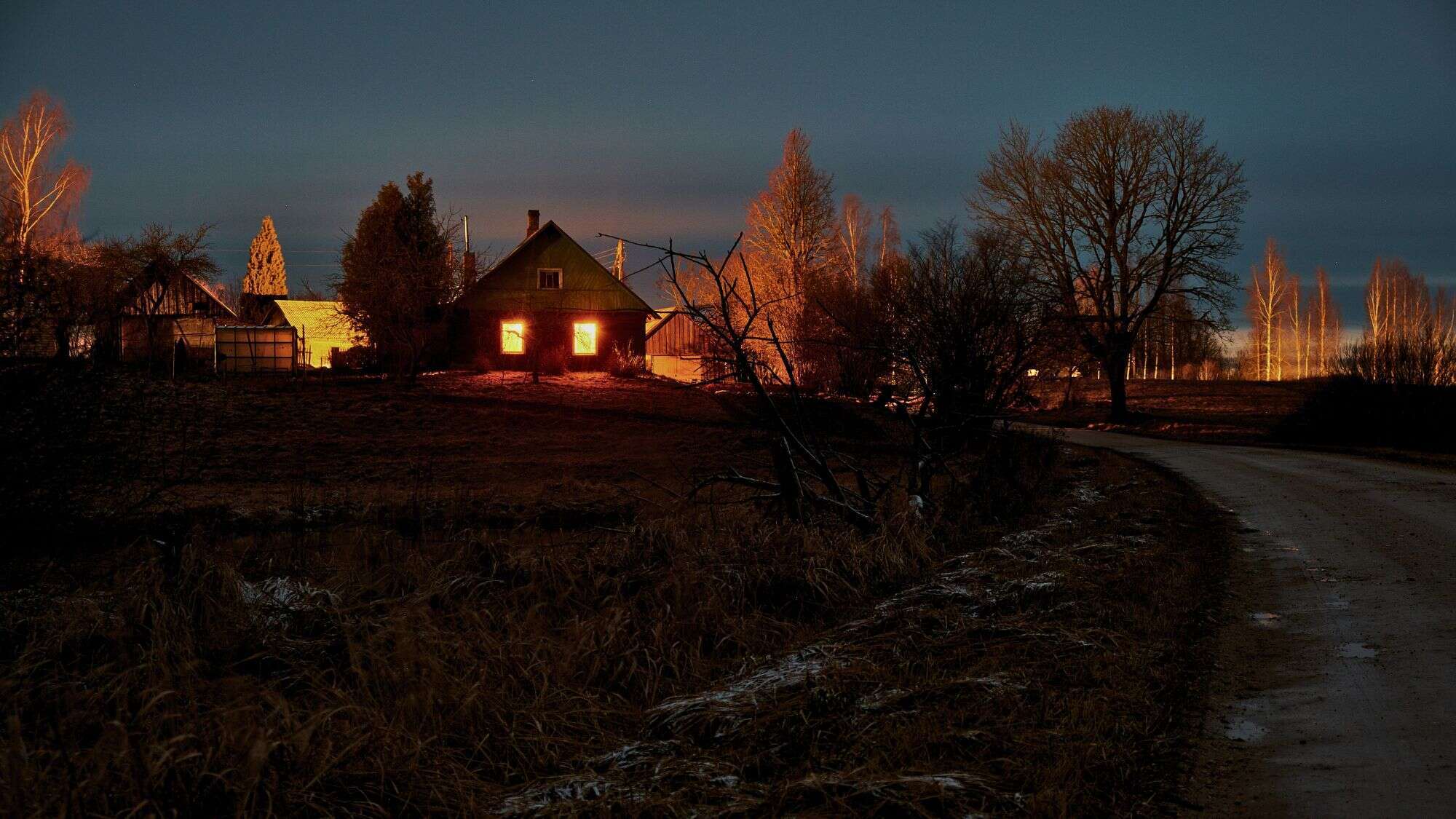 A dark country cabin radiates an ominous orange glow through its windows, surrounded by bare trees and a dirt road under a twilight sky.