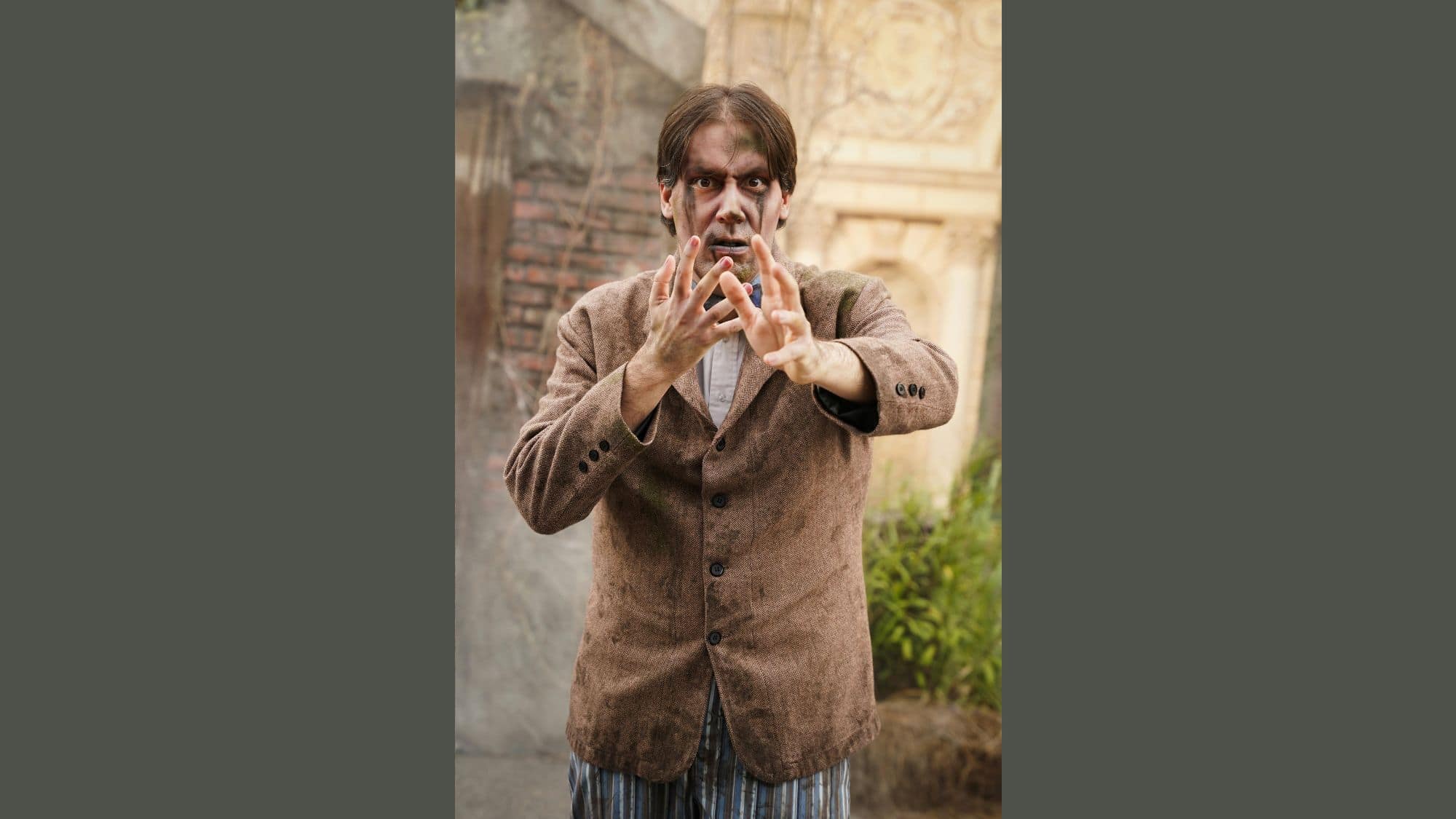 A man dressed as a zombie with smeared makeup, disheveled hair, and ragged clothing poses mid-motion in front of a foggy, stone wall backdrop.