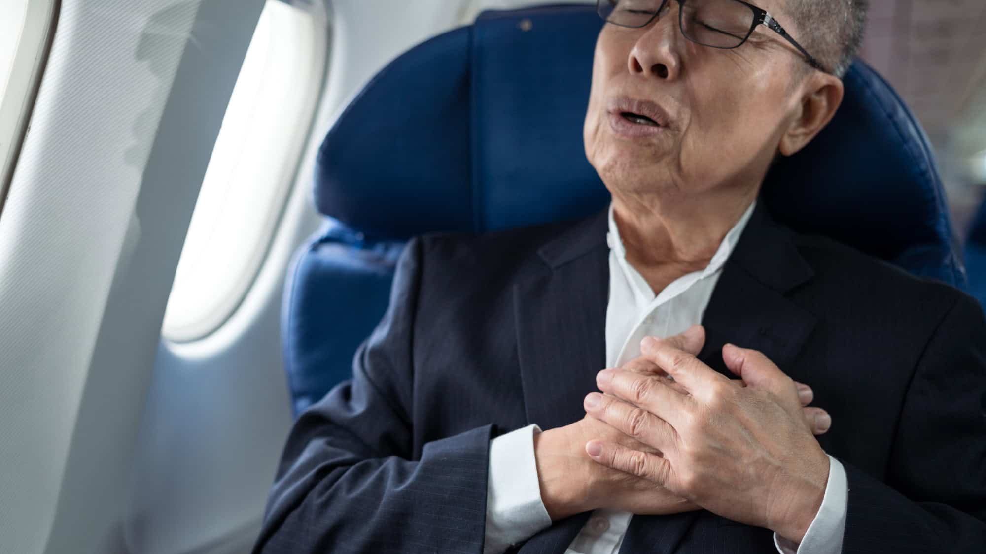 An older man in a suit clutches his chest with a pained expression while seated on an airplane.