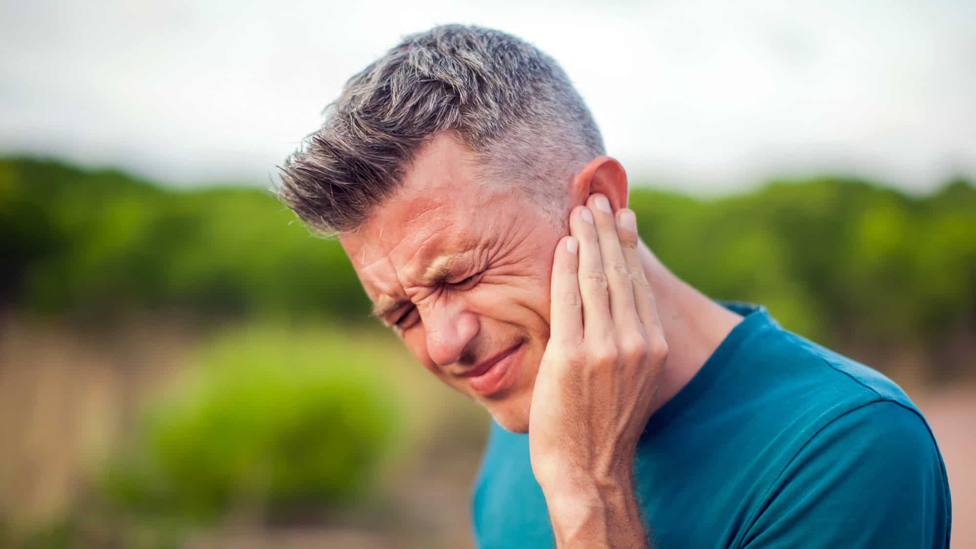 A man outdoors presses his hand to his ear with his eyes closed tightly, showing signs of discomfort.