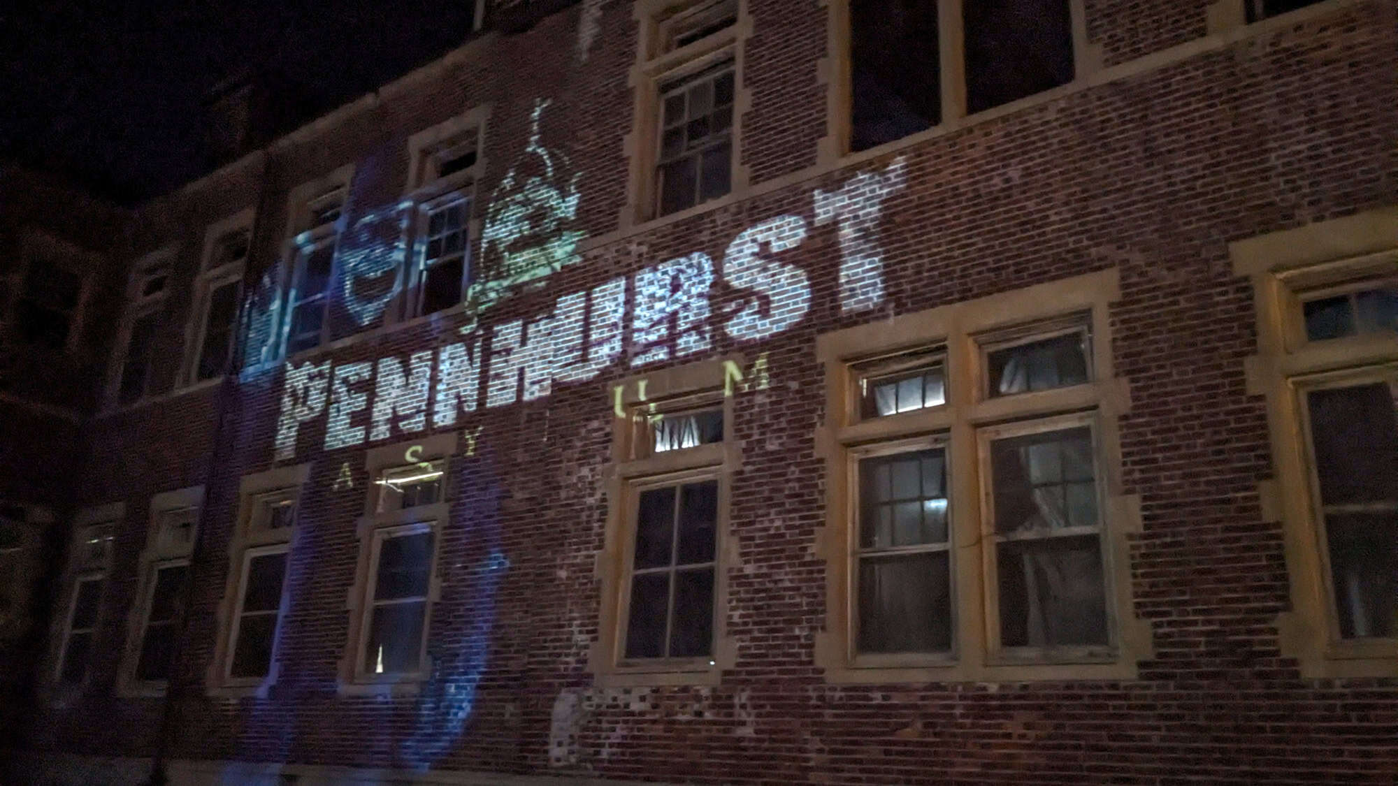 The words "Pennhurst Asylum" and eerie faces are projected in glowing white light across the brick façade of a historic building at night.