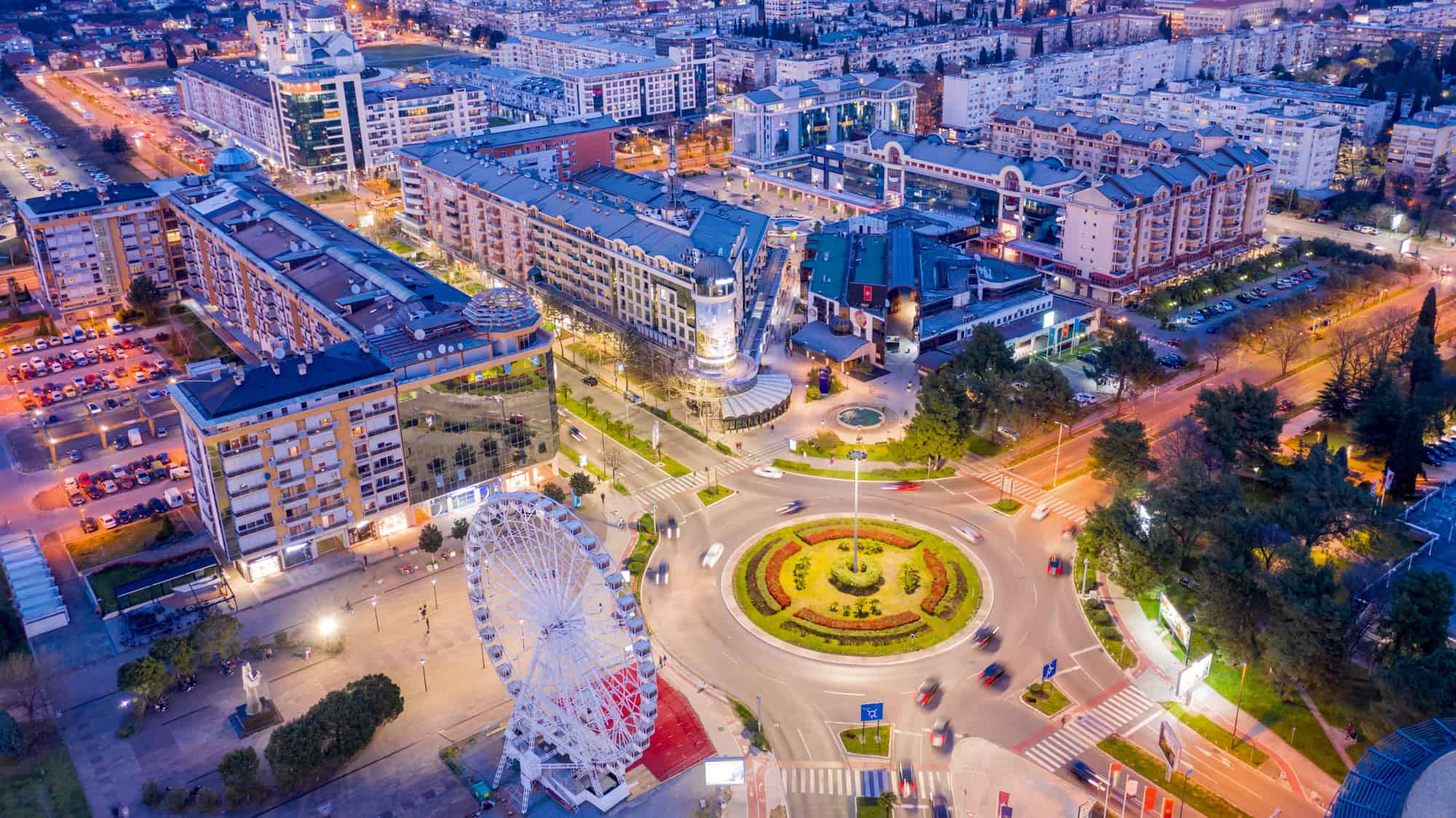 An aerial evening view of Podgorica, Montenegro shows a traffic roundabout with flower beds and a Ferris wheel surrounded by modern buildings lit up at dusk.