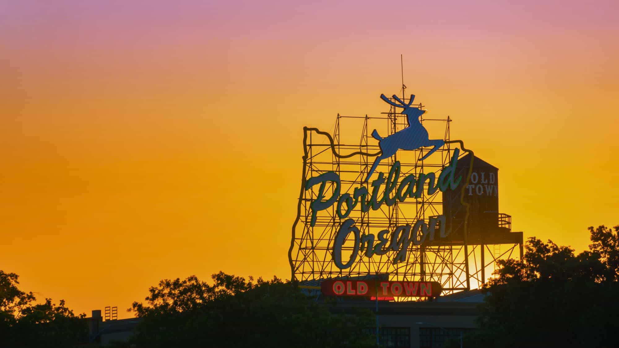 The iconic Portland Oregon neon sign featuring a leaping stag is silhouetted against a vivid orange and purple sunset sky, with "Old Town" glowing below.