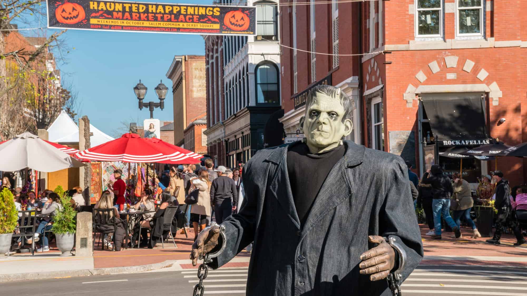 A person dressed as Frankenstein stands in front of a crowd and vendor booths under a “Haunted Happenings Marketplace” banner during a festive Halloween event in Salem, Massachusetts.