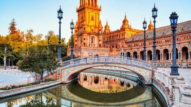 A richly tiled footbridge arches over a calm canal in front of the ornate brick towers and colonnades of Plaza de España in Seville, glowing warmly in golden hour light.