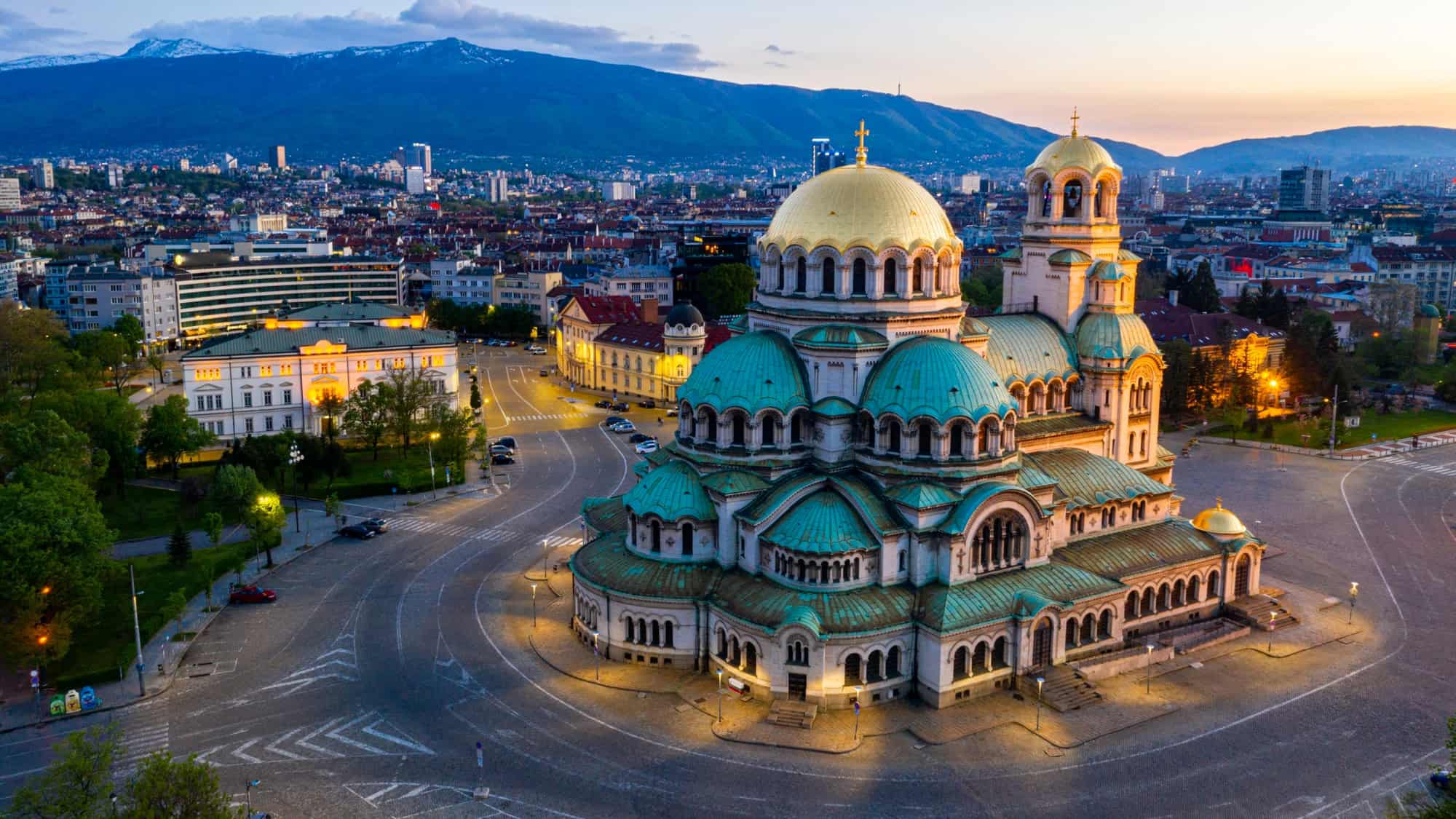 The iconic green and gold domes of Alexander Nevsky Cathedral in Sofia, Bulgaria glow at sunset, with the city and mountains stretching out behind.