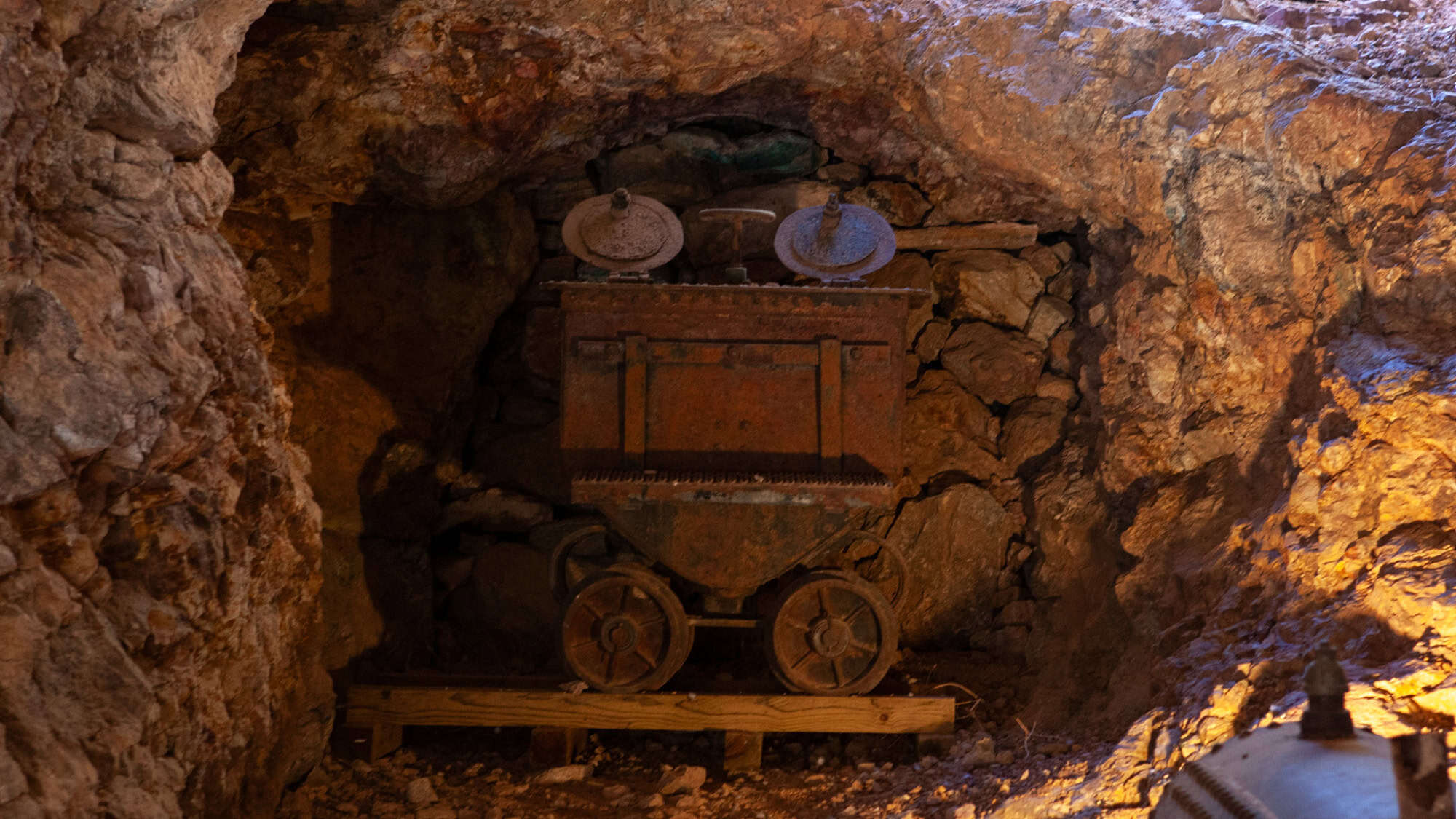 A rusty ore cart sits on rails inside a dimly lit gold mine tunnel, surrounded by jagged rock walls and a backdrop of stacked stones.