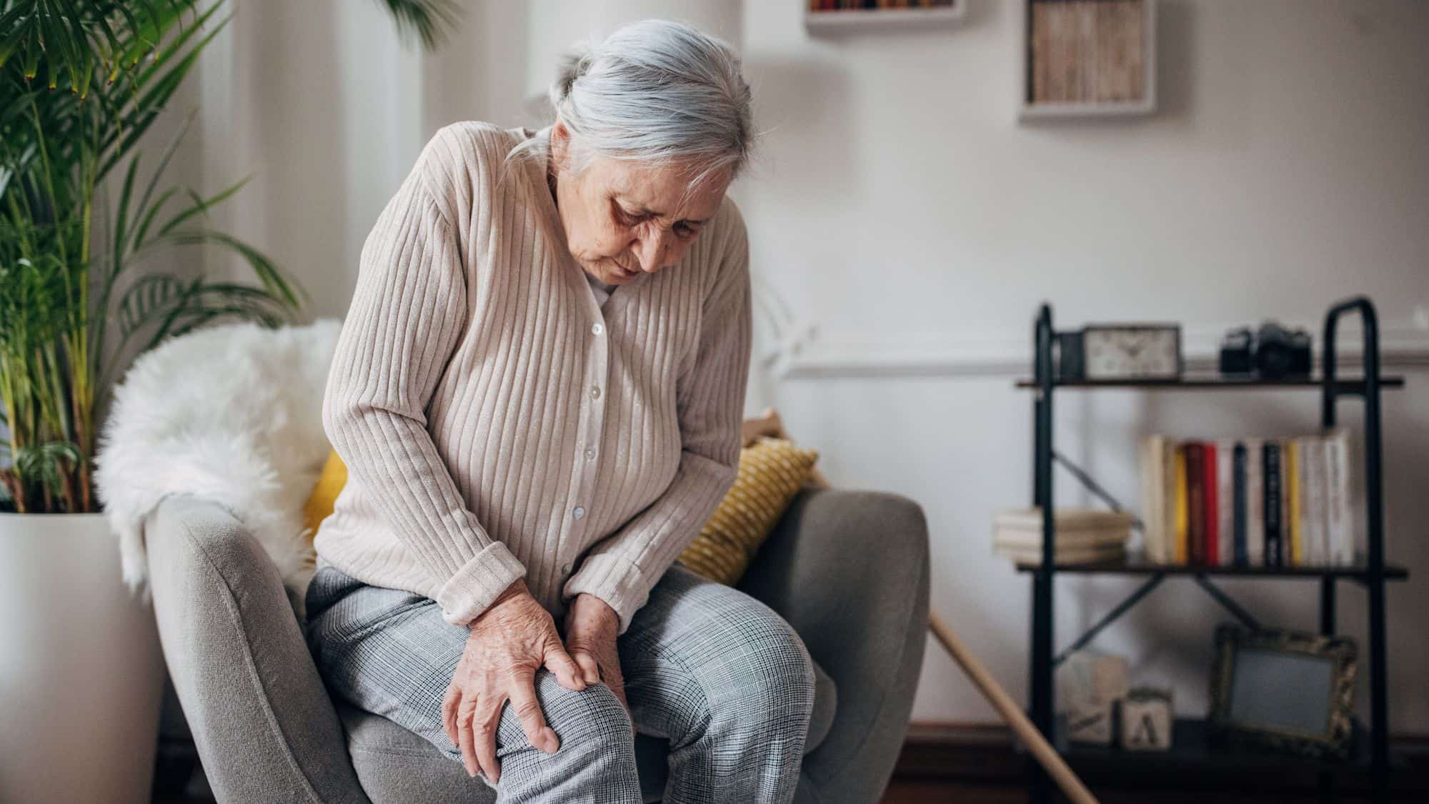 An elderly woman sitting in a chair clutches her knee with a pained expression, suggesting joint discomfort.