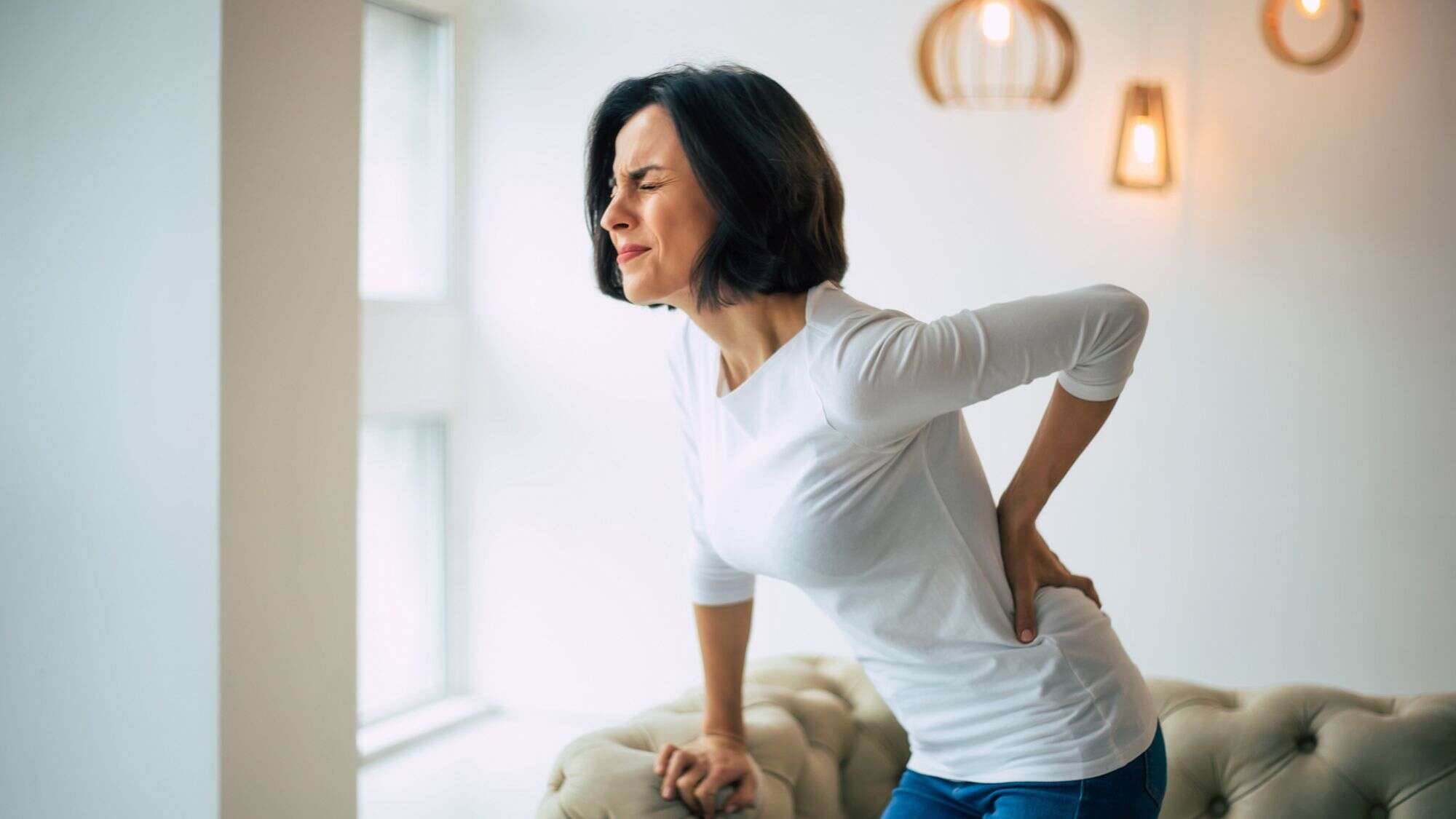 A woman in a white shirt leans forward with a strained face, pressing her hand against her lower back in pain.