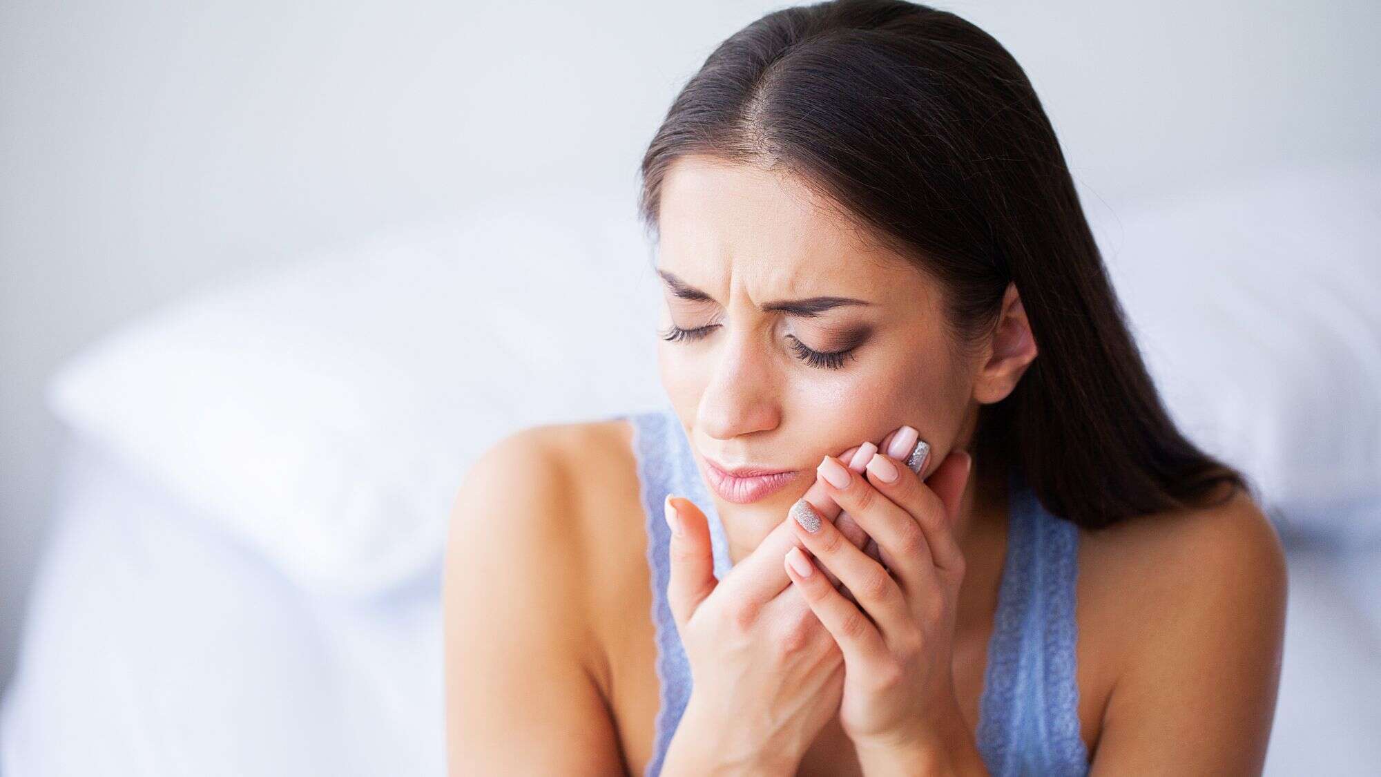 A woman in a blue tank top grimaces and holds her jaw, indicating tooth pain.