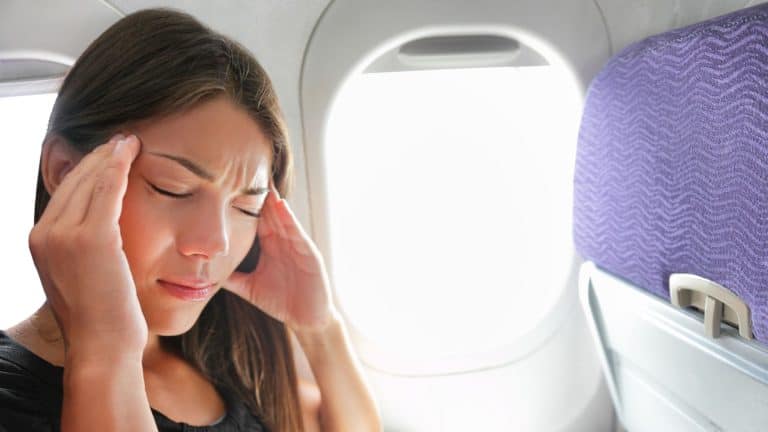 A woman seated by a plane window presses her temples with her eyes closed, suffering from a headache.