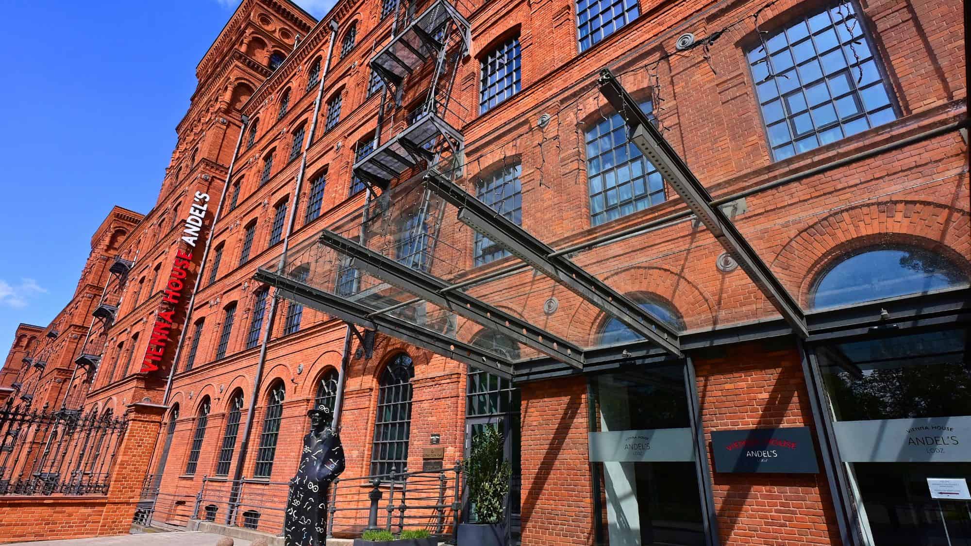 The red-brick industrial-style building of Vienna House Andel’s in Łódź, Poland features large black-trimmed windows, metal fire escapes, and a modern glass awning over the entrance.
