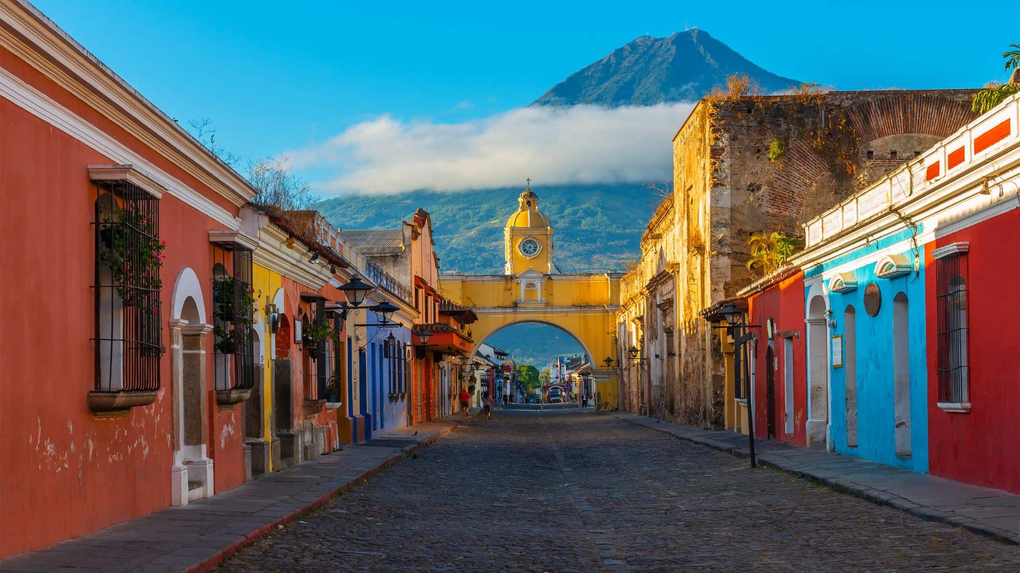 A cobblestone street in Antigua, Guatemala, leads to the iconic yellow Santa Catalina Arch, with a misty volcano rising in the background.