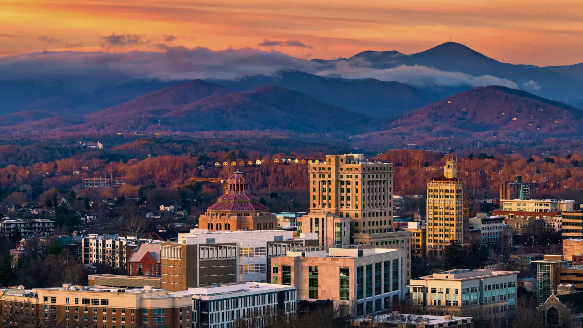 The cityscape of Asheville, North Carolina glows under a warm sunset, with the Blue Ridge Mountains rising misty and layered in the background.