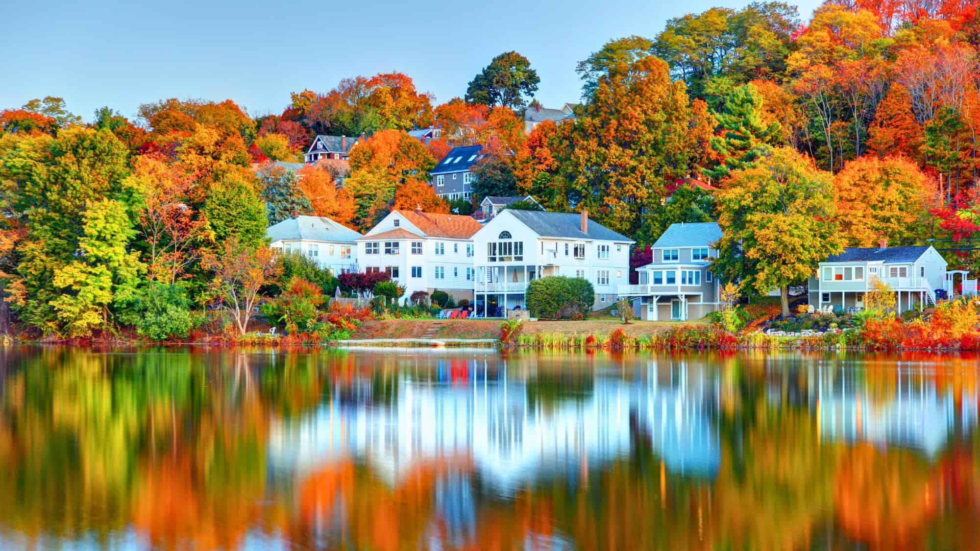 White and pastel-colored homes sit along a calm lake, where brilliant fall trees reflect vividly in the water.
