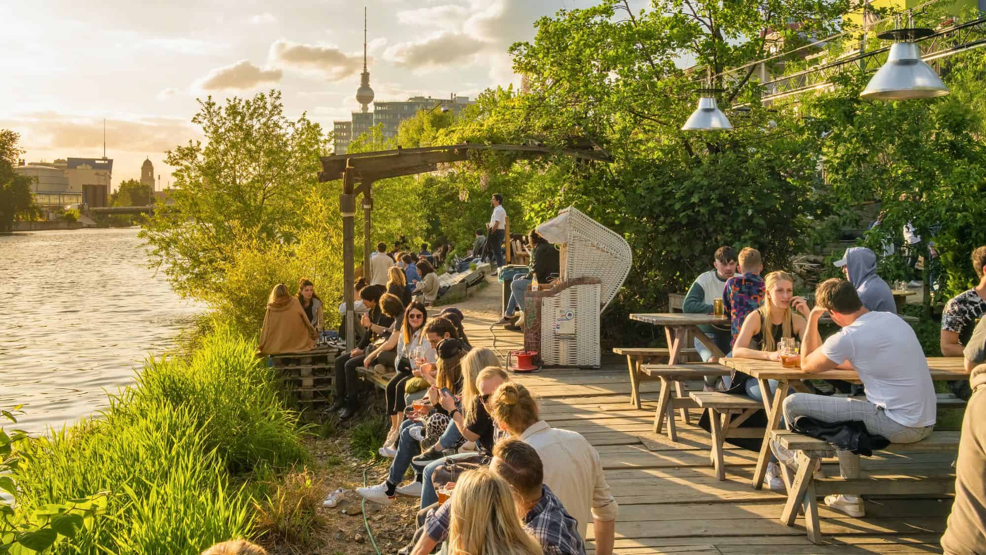 People relax at a lively riverside wooden deck bar in Berlin, with the iconic TV Tower visible in the sunset-lit background.