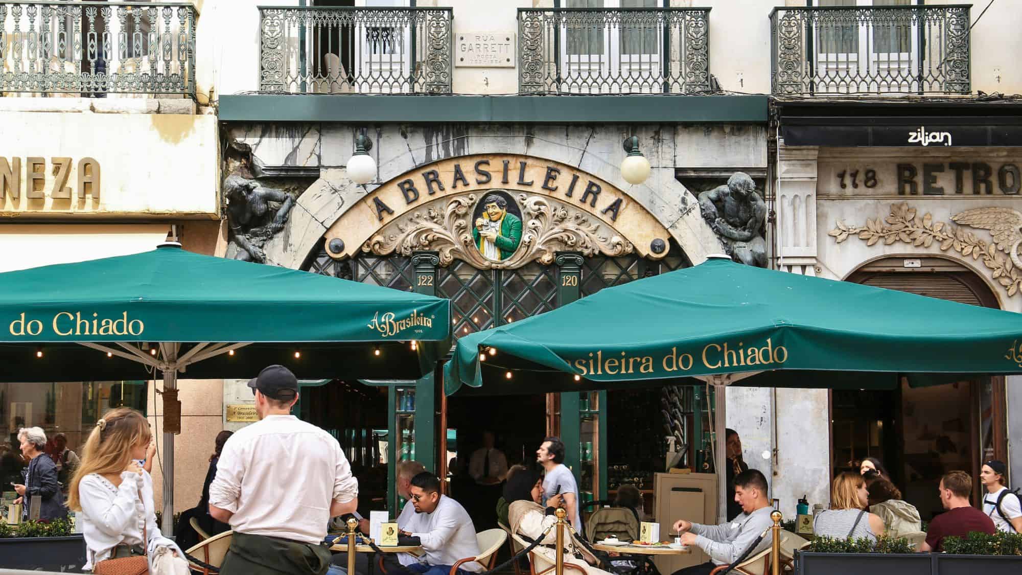 An ornate historic café with green awnings and sculptures flanking the entrance serves patrons at outdoor tables in Lisbon's Chiado district.