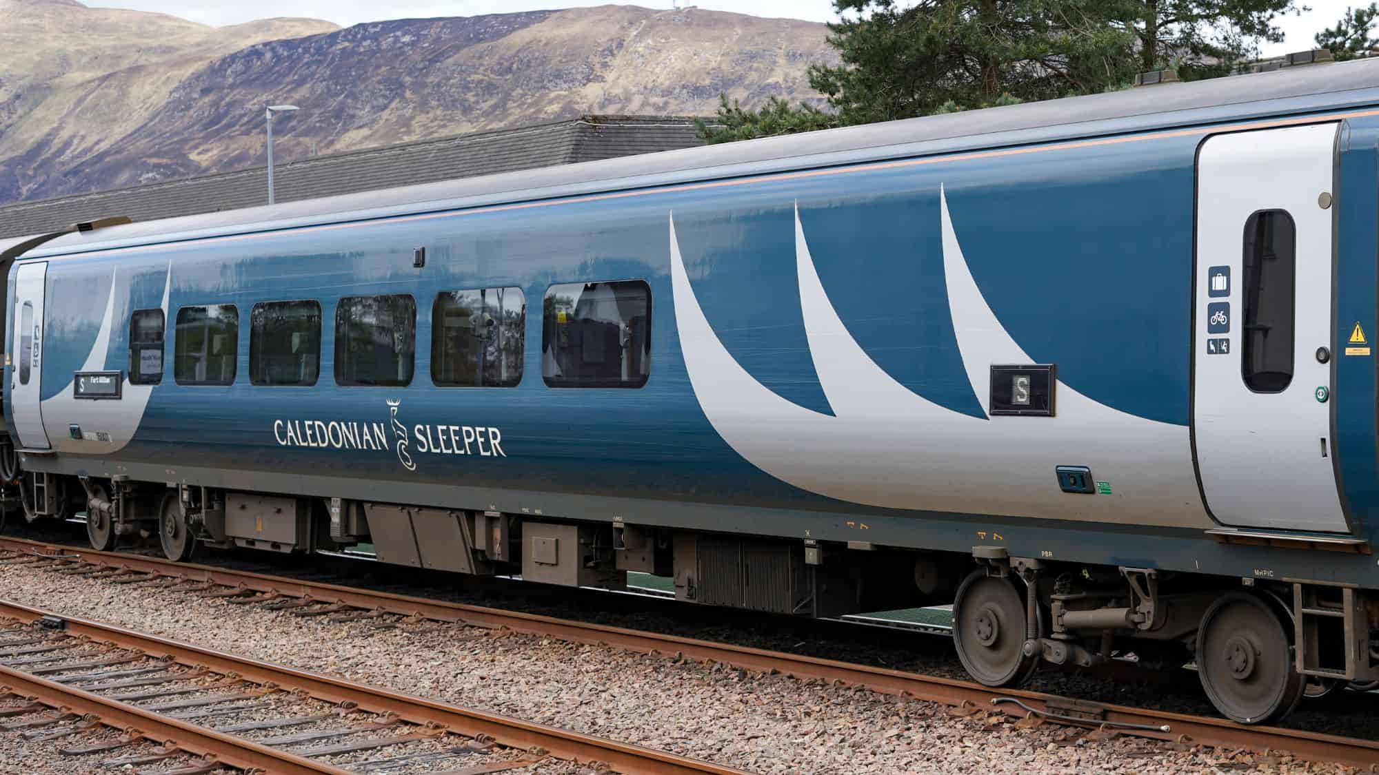 A blue and silver Caledonian Sleeper train car sits on the tracks in front of a hilly Scottish backdrop, with bold white branding and symbols indicating luggage, bike, and accessibility.