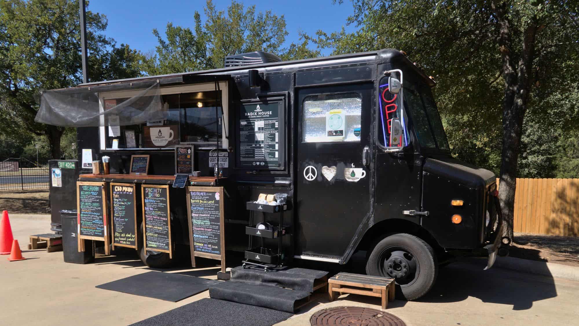 A sleek black mobile coffee truck named Radix House offers specialty drinks from a curbside setup under trees in a sunny suburban area.