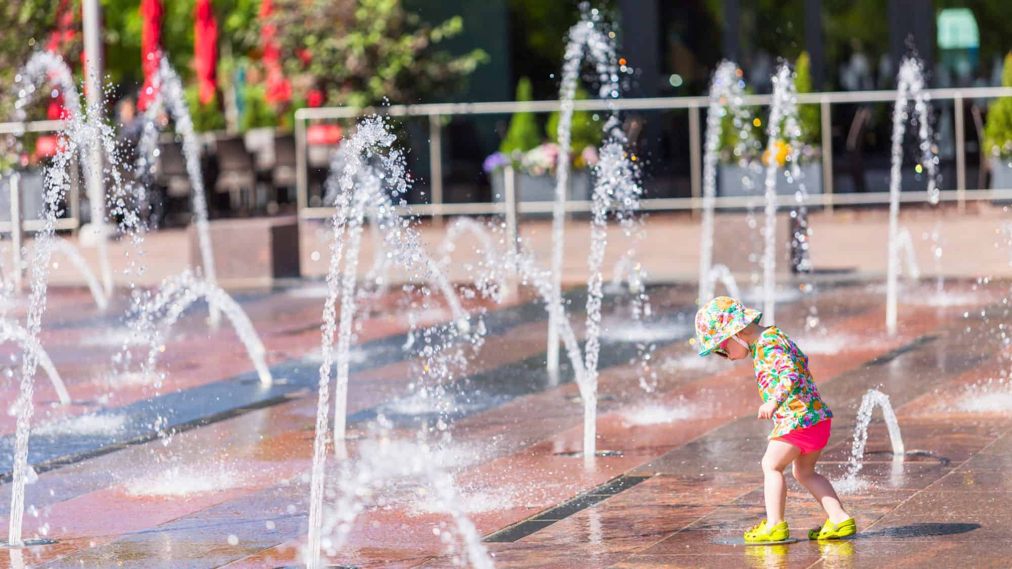 A young child in bright clothes and a sunhat joyfully plays among spouting water jets in a public splash pad on a sunny day.
