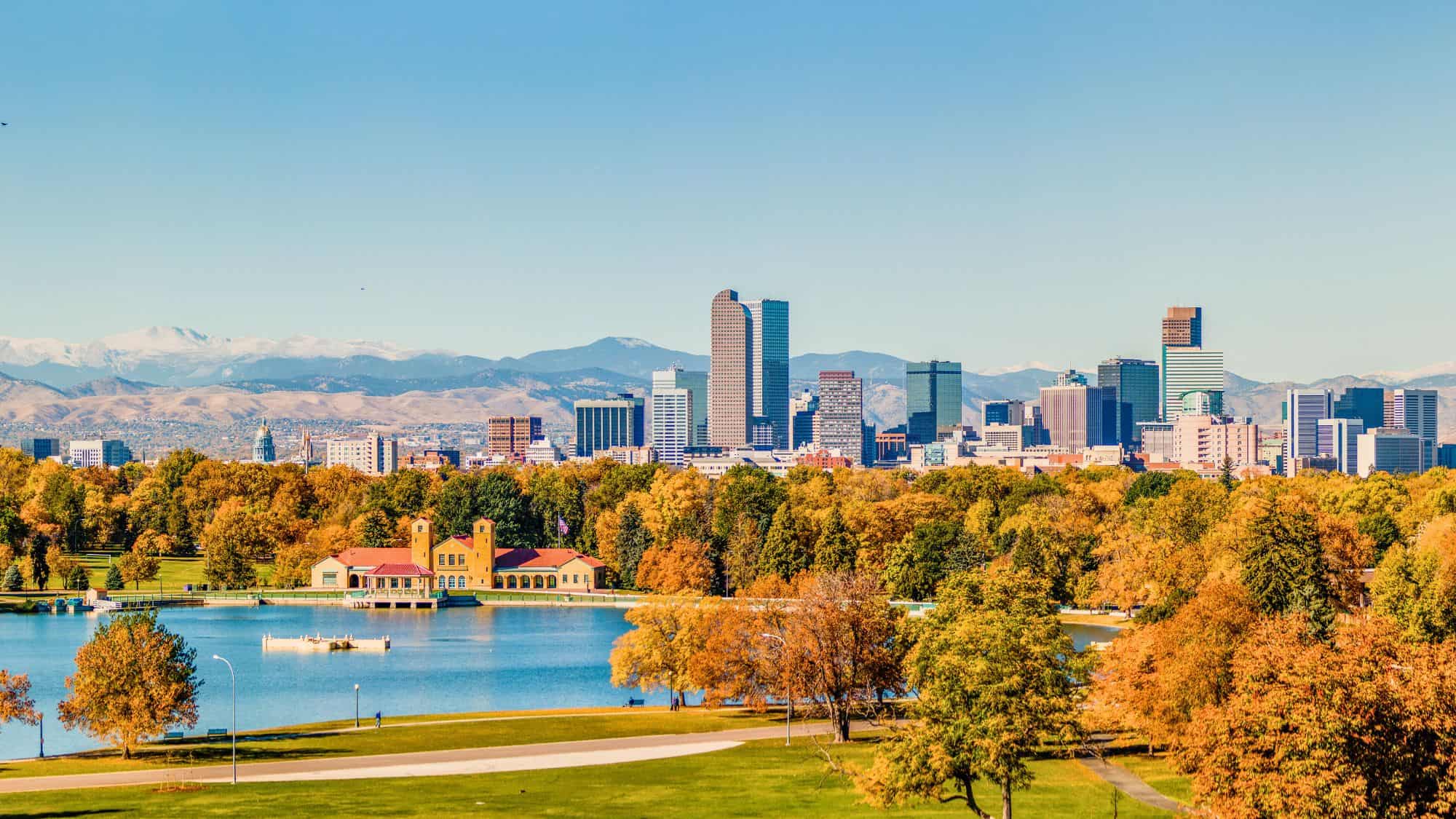 The Denver skyline rises behind a vibrant canopy of fall foliage, with snow-capped Rocky Mountains and a blue lake in the foreground creating a layered scenic view.