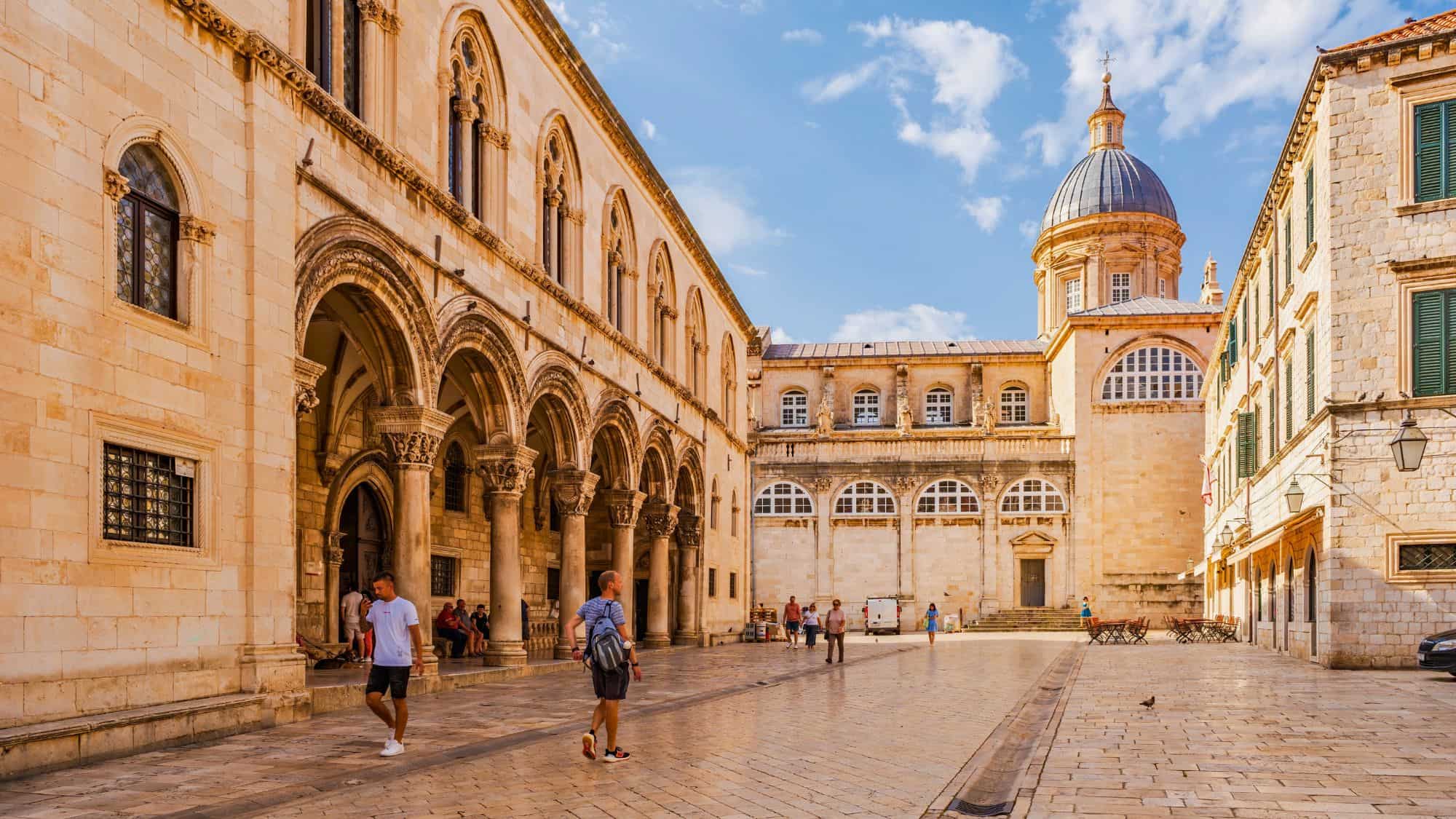 Tourists walk through a historic courtyard in Dubrovnik, Croatia, surrounded by grand arched colonnades and a domed cathedral glowing in the warm afternoon sun.