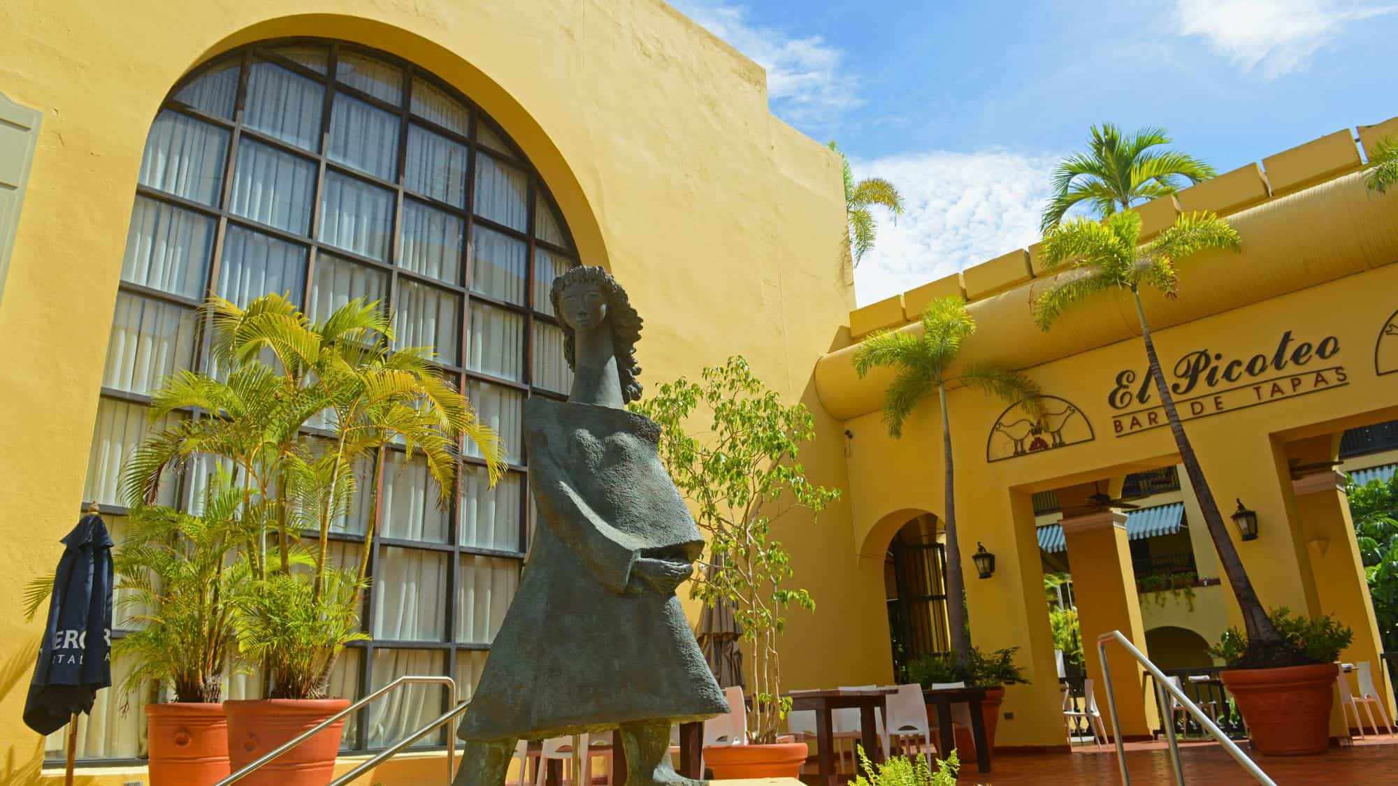 A bronze sculpture stands outside the yellow stucco building of El Picoteo Bar de Tapas, surrounded by potted palm trees and patio seating under a sunny sky.