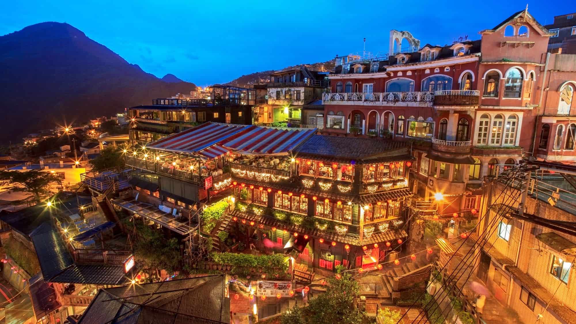 A hillside town in Taiwan glows at night with colorful lanterns, lively restaurants, and ornate architecture under a deep blue sky.
