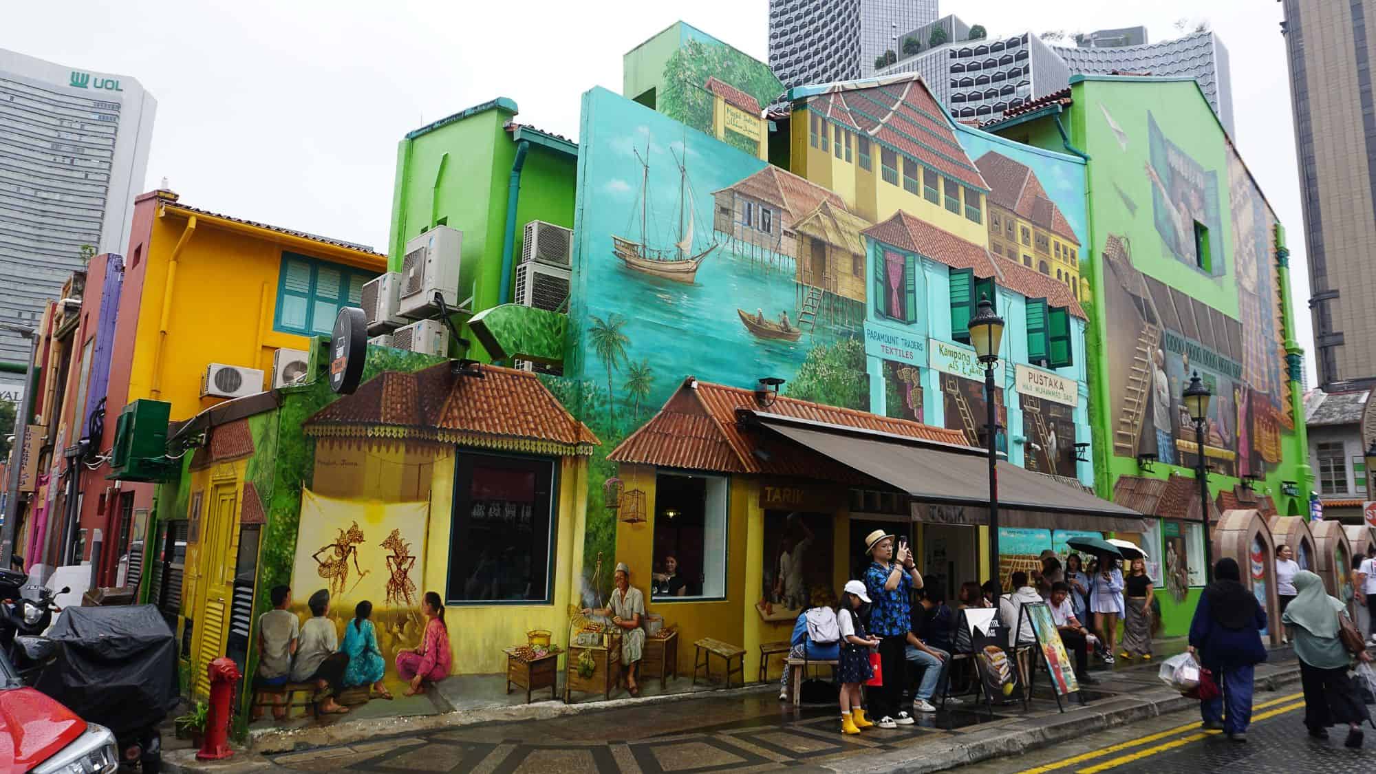 A vibrant street in Singapore features a café set against colorful murals depicting village life, attracting passersby with its eclectic charm.