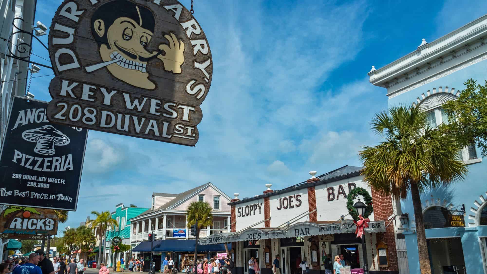 A lively scene on Duval Street in Key West features iconic signage for Sloppy Joe’s Bar and Durty Harry’s, with palm trees and colorful buildings lining the bustling street.